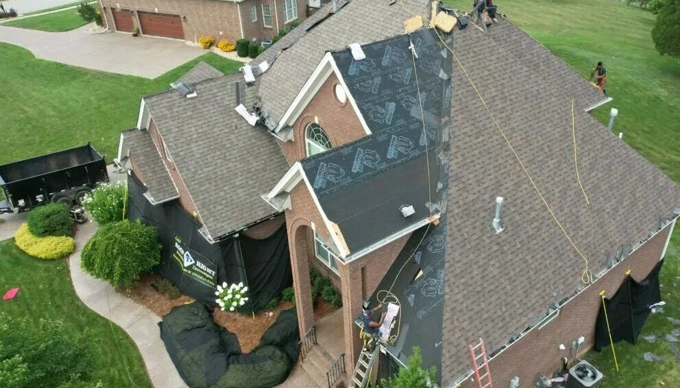 A house undergoing roof repairs with workers installing new roofing materials and black underlayment on the roof.