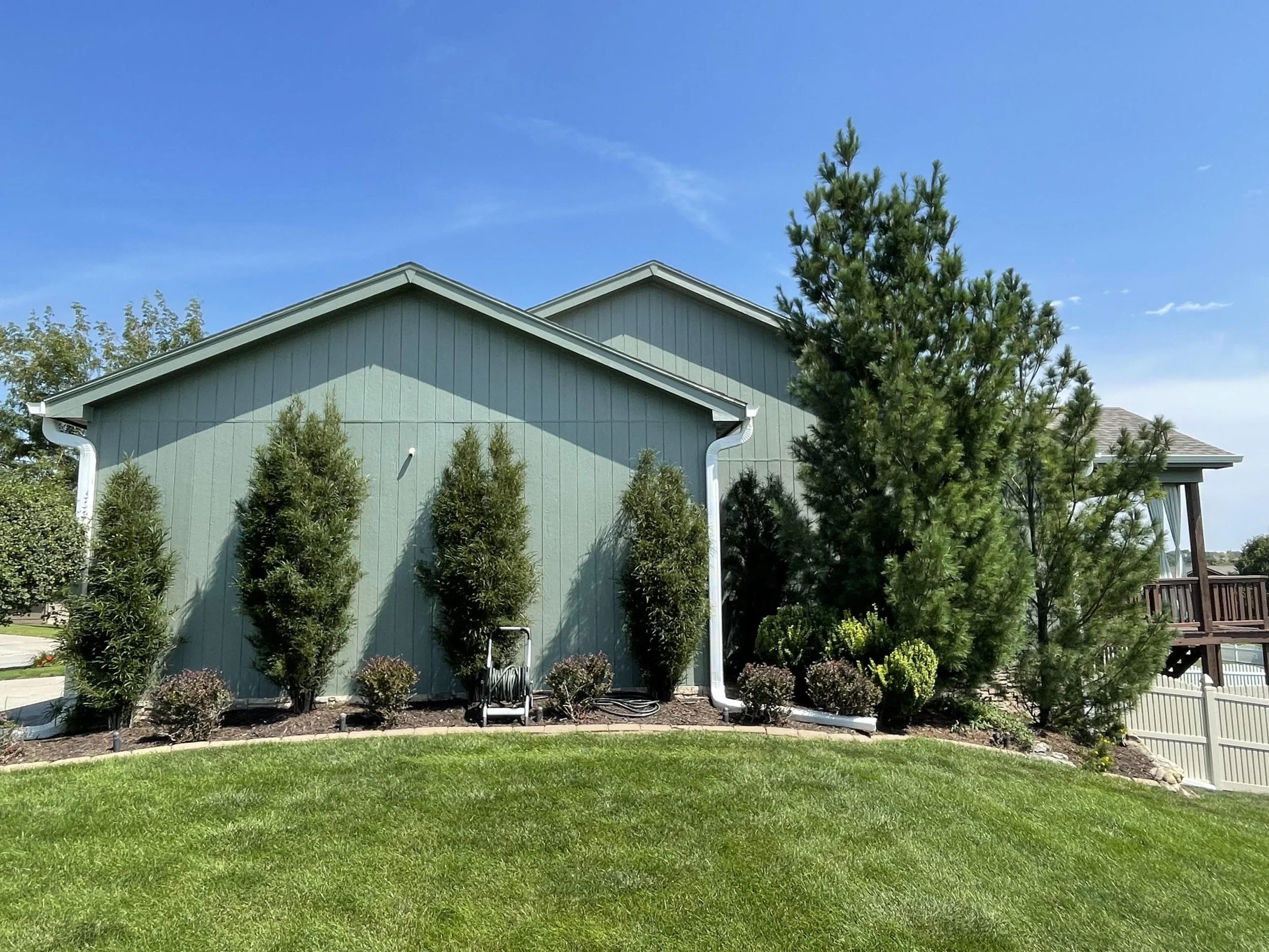 A two-story house with gray siding and white trim, surrounded by a well-maintained garden with various bushes and plants, under a clear blue sky.