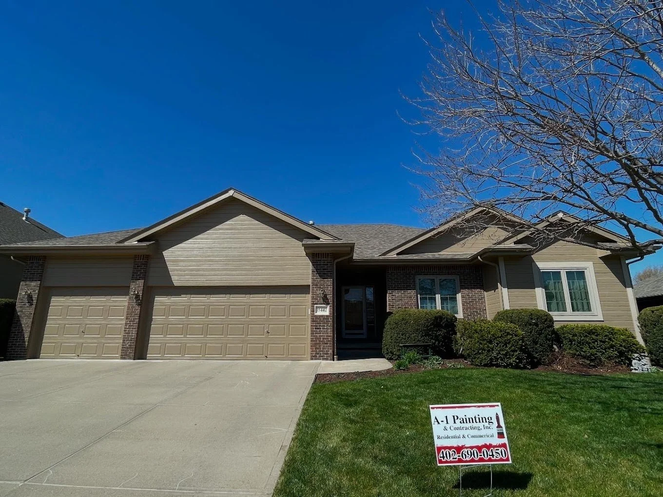 A two-story suburban house with a three-car garage, tan siding, brick accents, a front porch, and a lawn with bushes. There is a sign in the lawn advertising painting and contracting services with contact phone number 402-690-0450.