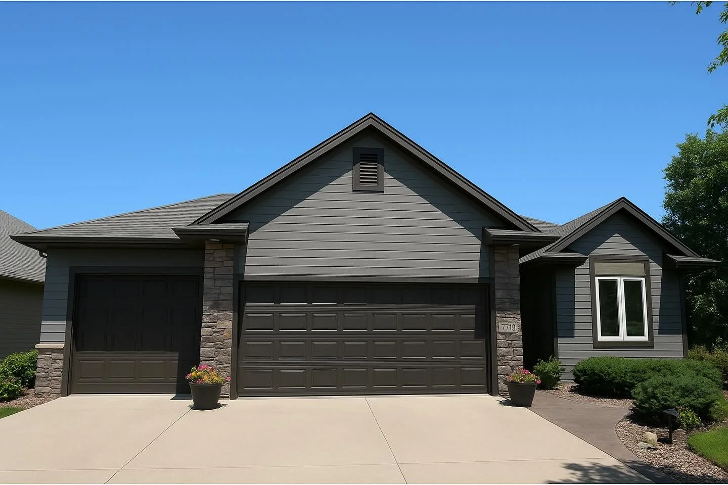 A modern two-story house with dark gray siding and stone accents, a three-car garage, and landscaping with potted plants and bushes under a clear blue sky.