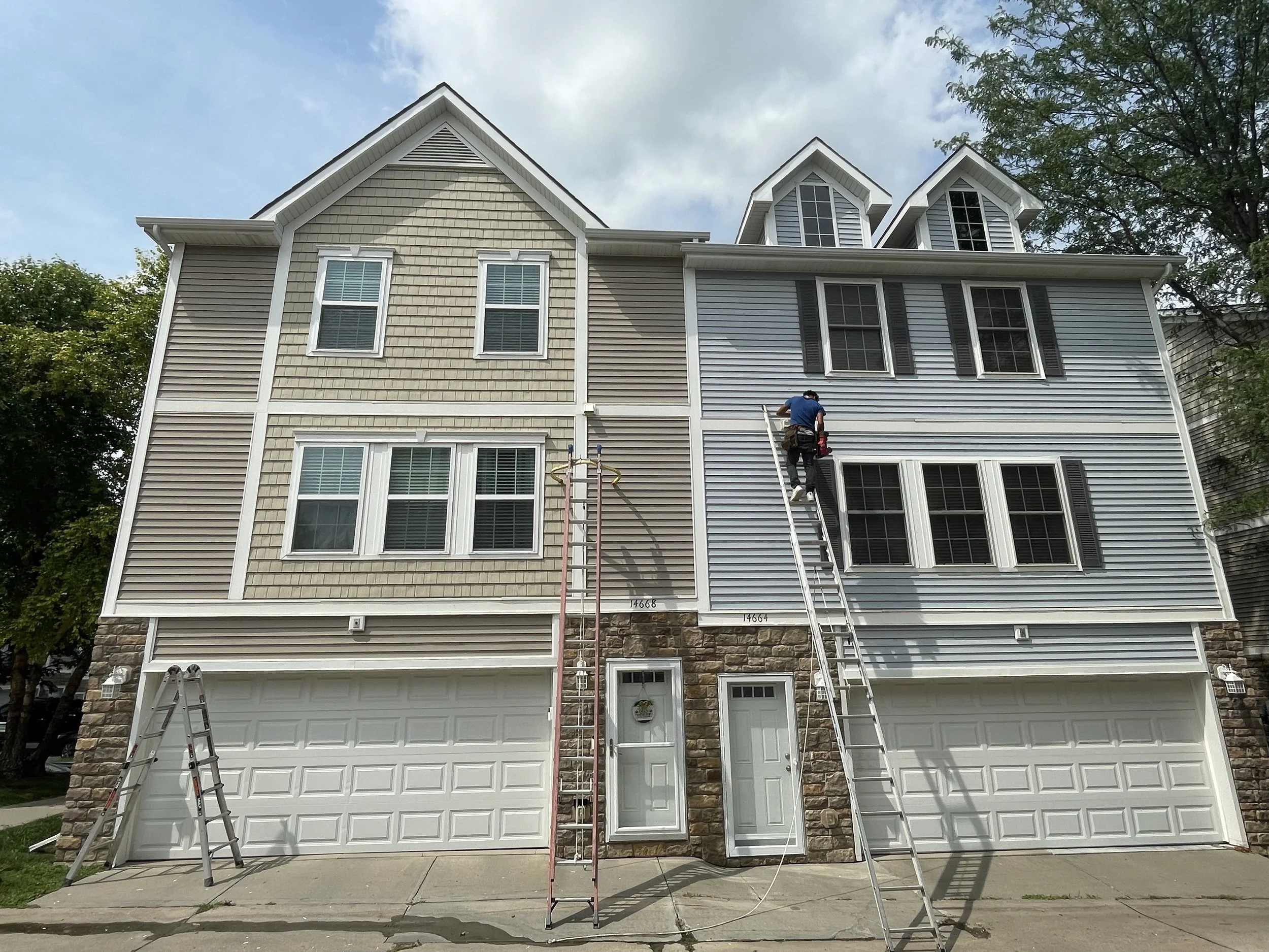 A multi-story house with a gable roof, siding in beige and gray, three garage doors, and a person working on a ladder at the upper part of the house.