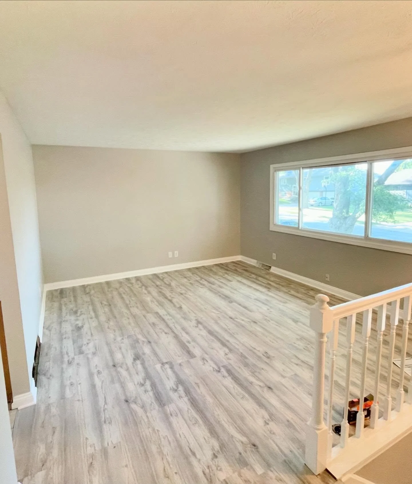 Empty living room with large window, light-colored walls, and wood-look flooring. Railing seen in the foreground.
