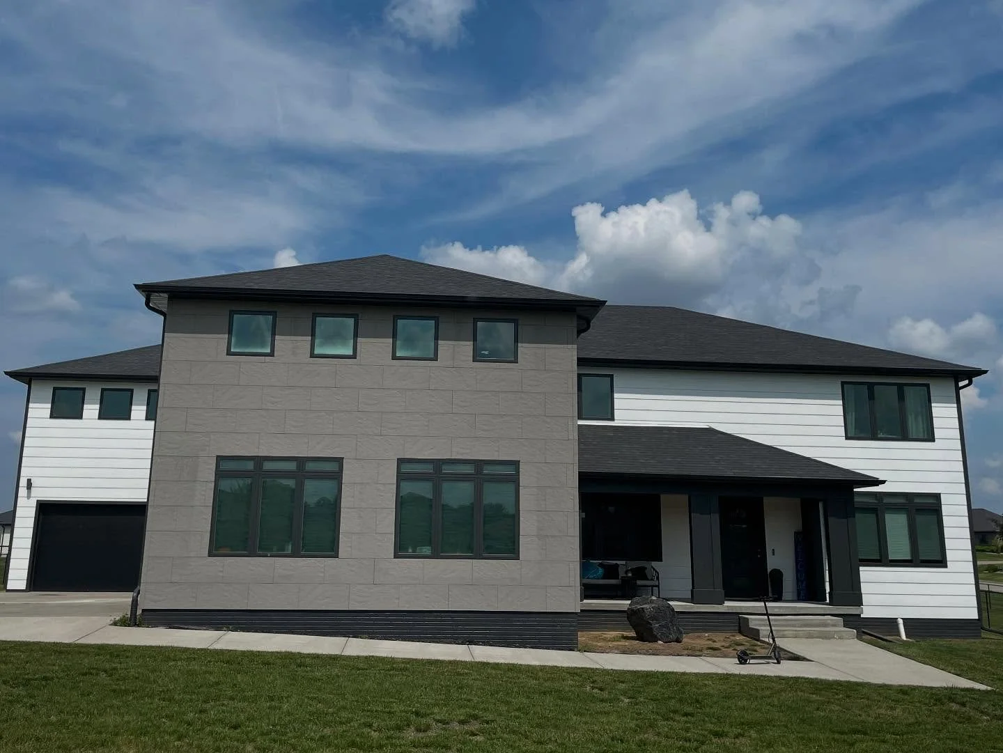Modern multi-story house with black roof, gray and white exterior walls, large windows, front porch, and green lawn under a blue sky with clouds.