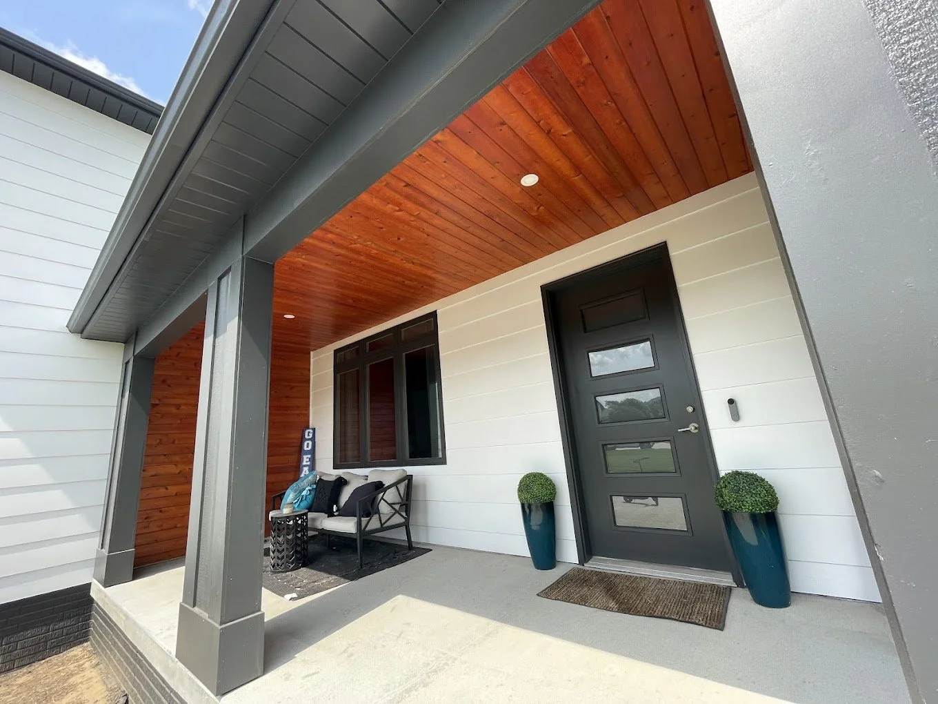 Front porch area of a house featuring a black front door with four rectangular windows, two large blue planters with round green bushes, a beige bench with cushions, a small black side table, and a welcome sign. The porch ceiling is wooden and the ho