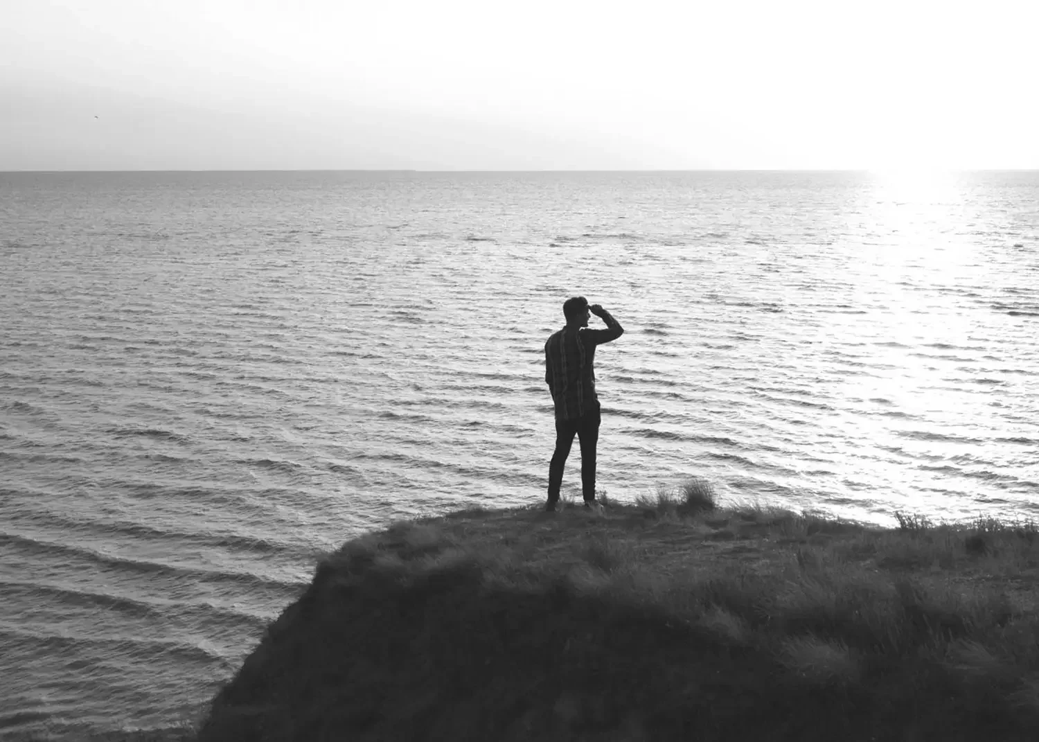A person standing on a grassy cliff overlooking the ocean, shielding their eyes from the sun, in black and white.