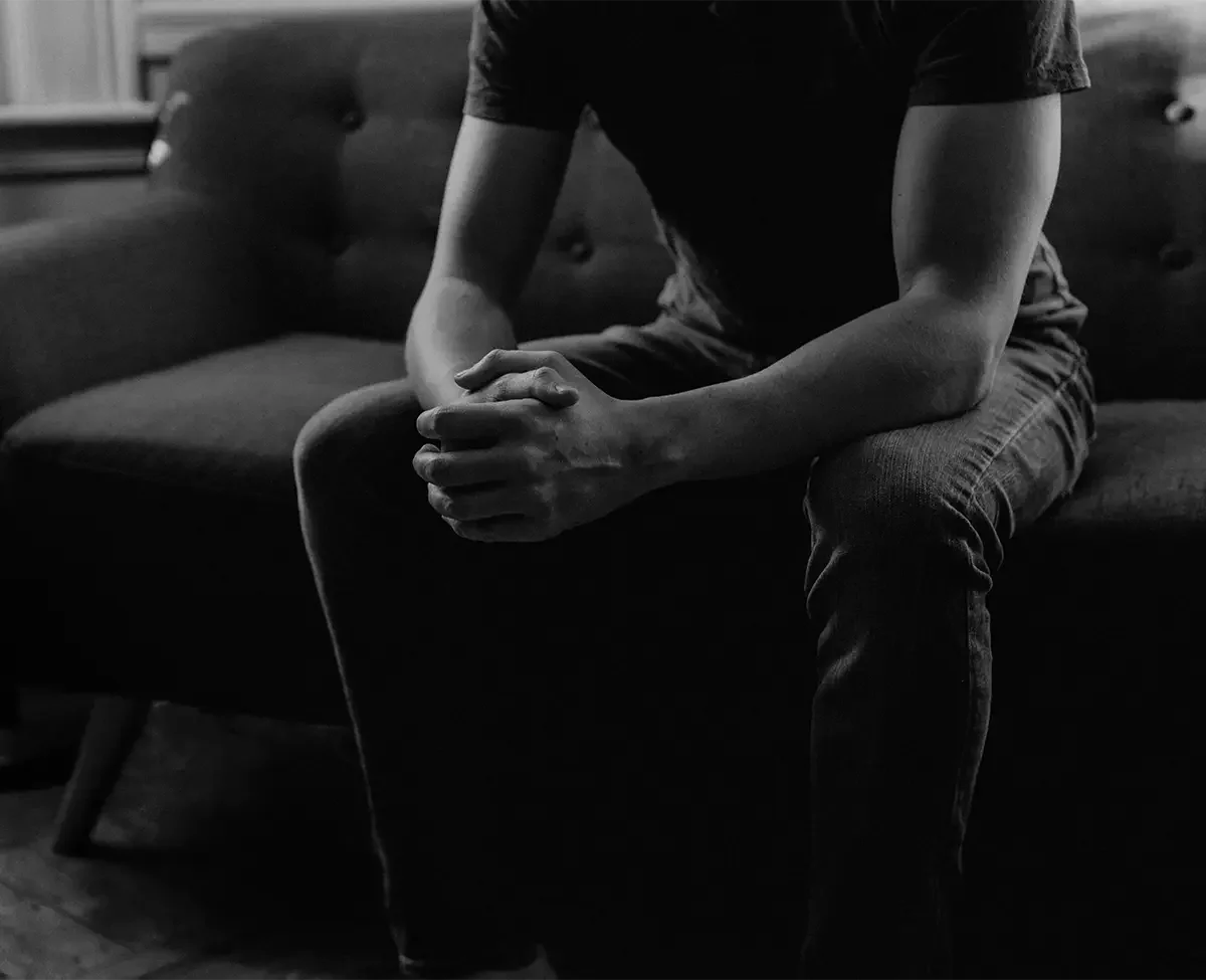 A person sitting on a sofa with hands clasped and head bowed, in a black and white photo.