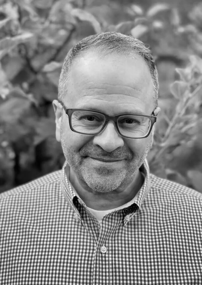 Black-and-white portrait of a middle-aged man wearing glasses and a checkered button-down shirt, smiling gently with leafy plants blurred in the background.