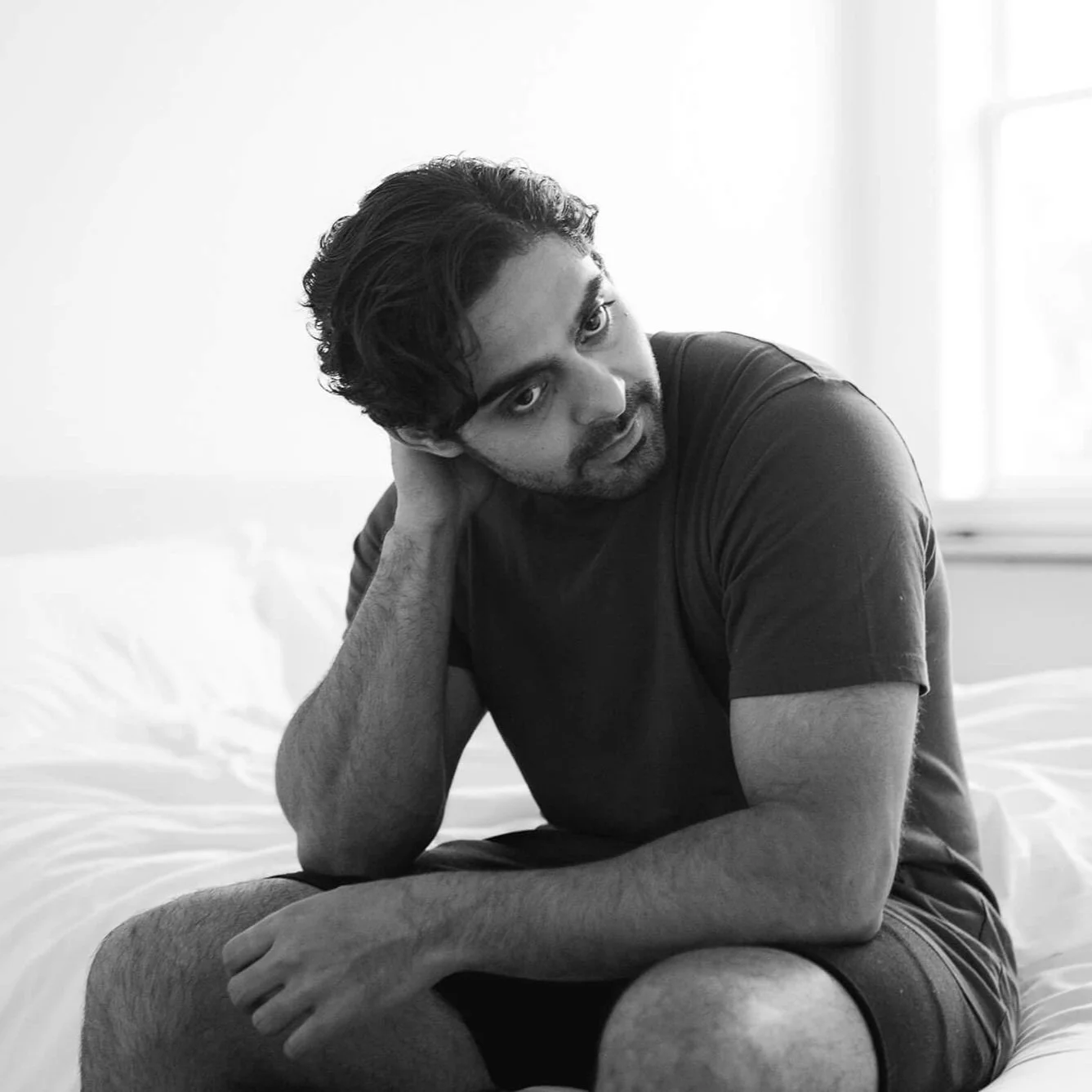 A man with dark hair and facial hair wearing a black shirt sits on a side of the bed and looks pensive and vulnerable