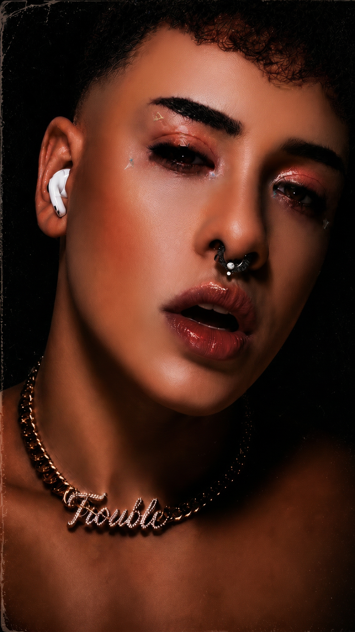 Close-up of a woman with short curly hair, wearing a white earbud, shiny lip gloss, a septum piercing, star-shaped face stickers, and a necklace that spells 'Trouble'.