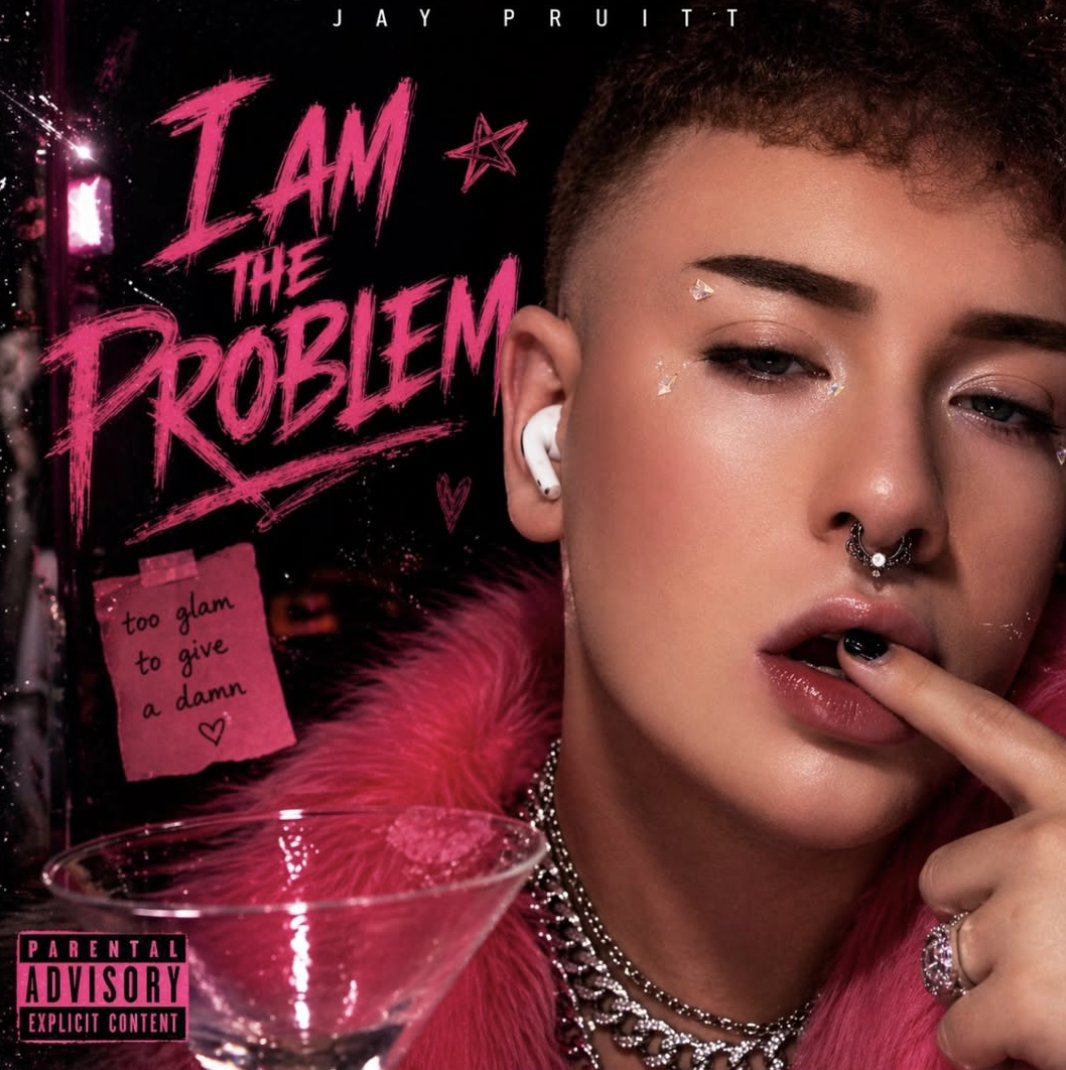Close-up of a person with short hair, wearing jewelry, makeup with glitter, and a pink fur coat, pointing to their lips. The background features a stylized album cover titled 'I AM THE PROBLEM' by Jay Pruitt with handwritten notes and stickers, including a parental advisory label.