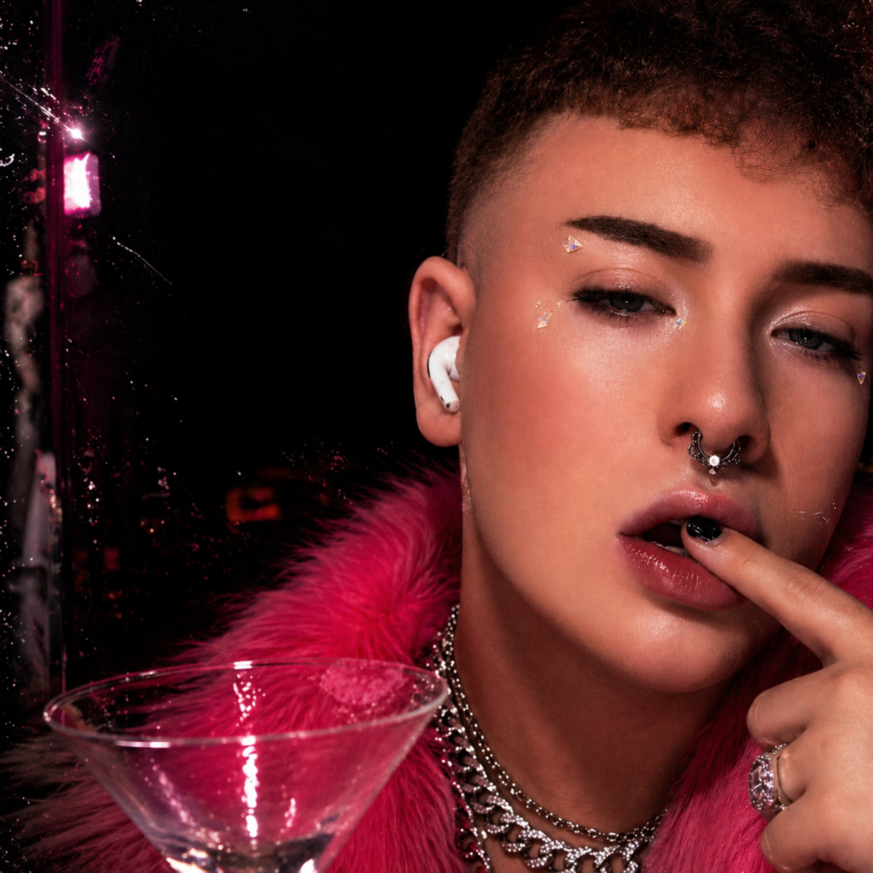 Close-up of a person with short hair, metallic makeup, and accessories, wearing a pink fur coat, with a cocktail glass in the foreground and a dark background.