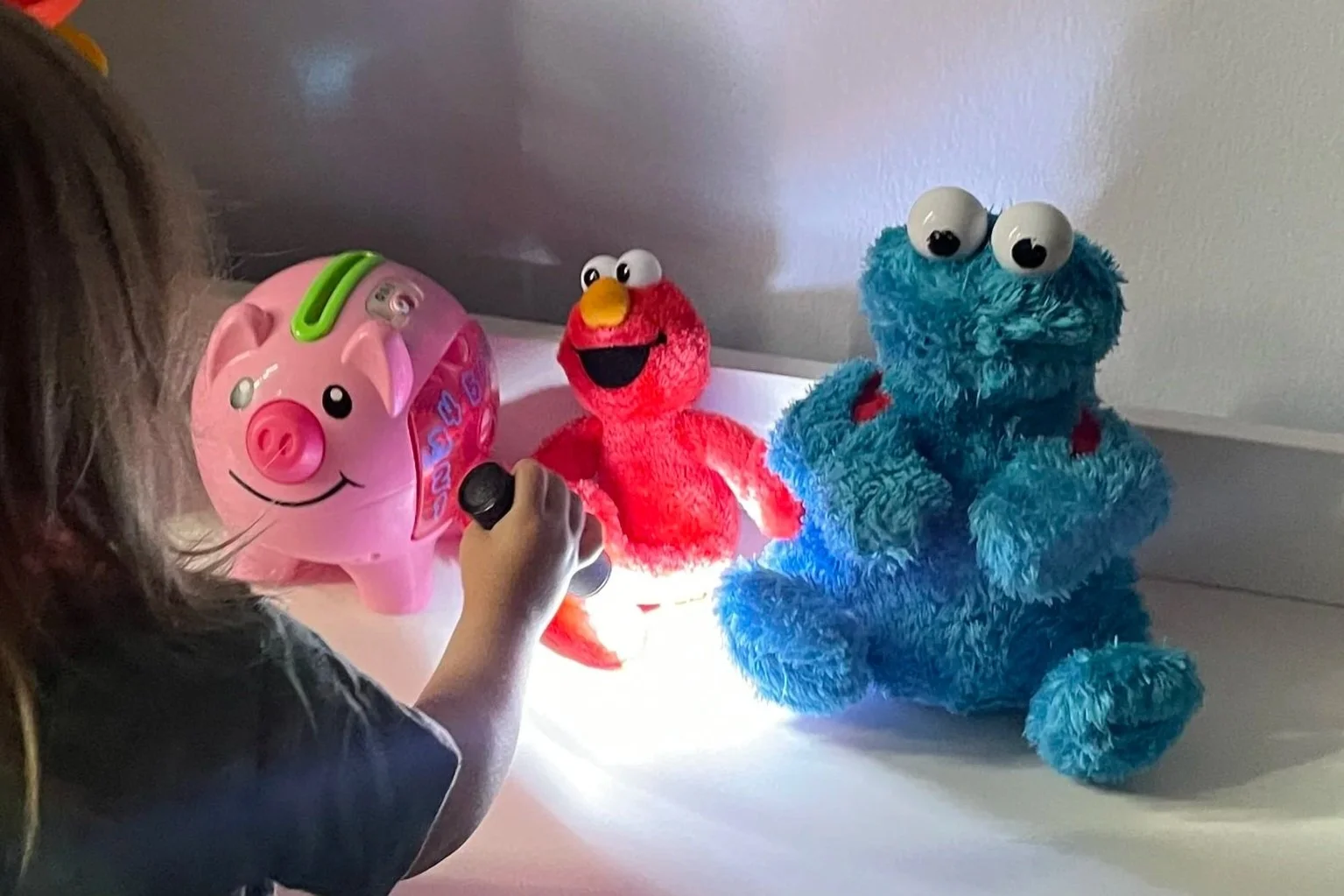 Child playing with plush toys and a piggy bank on a white surface, including a pink piggy bank, Elmo plush, and Cookie Monster plush.