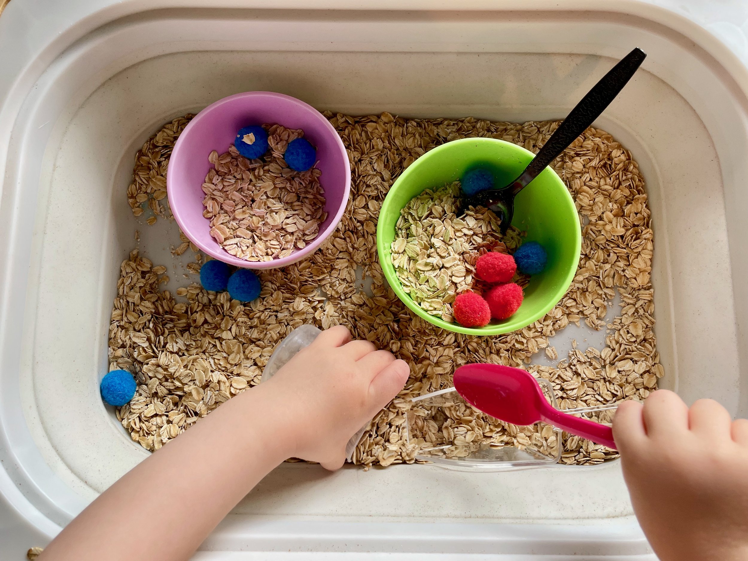 A child placing small colorful pom-poms into a container filled with oats, with other containers holding oats and more pom-poms nearby.