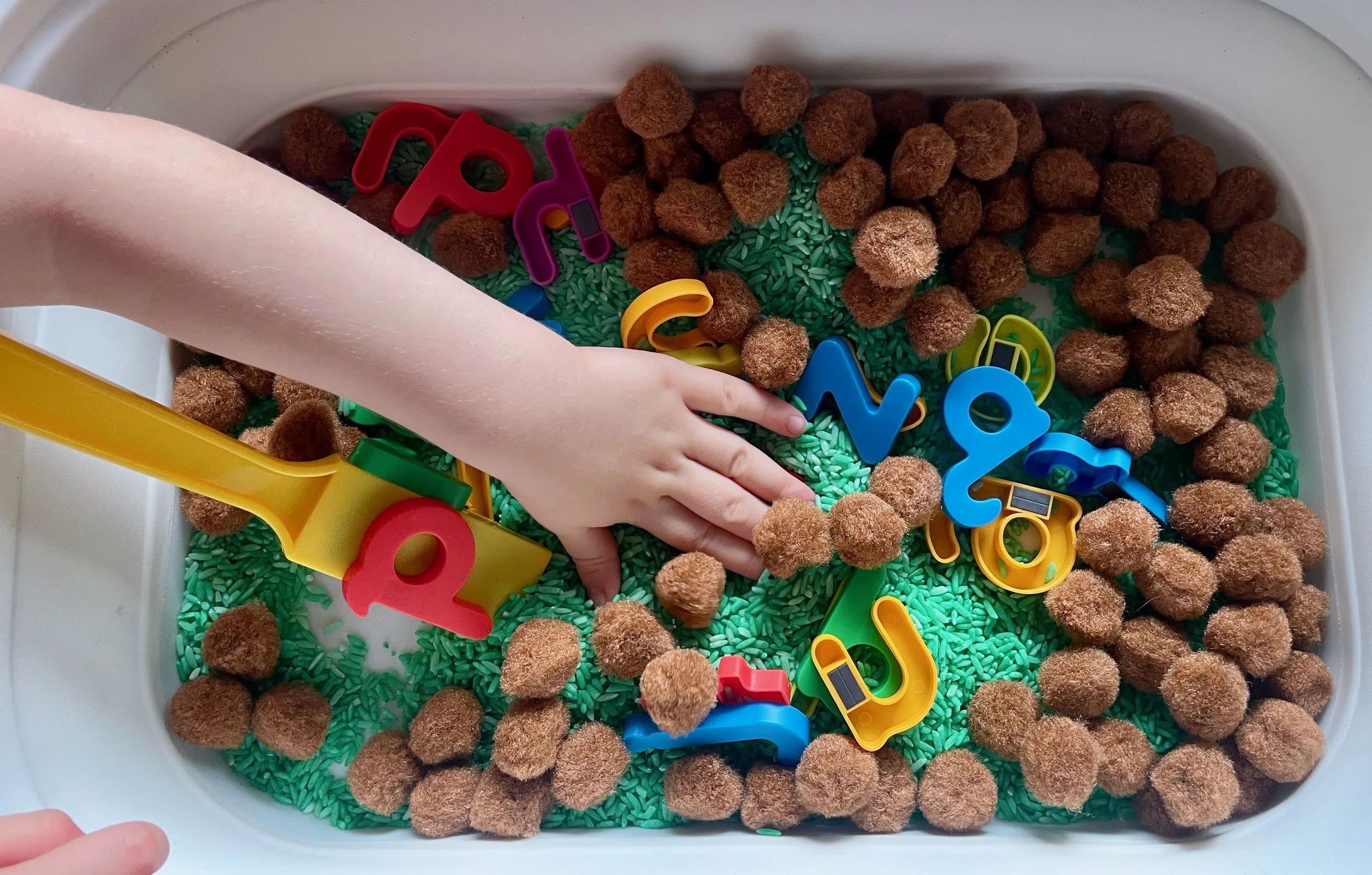 Toddler playing in a Chicka Chicka Boom Boom themed sensory bin with green rice, brown pom poms, magnetic letters, and a magnet wand.