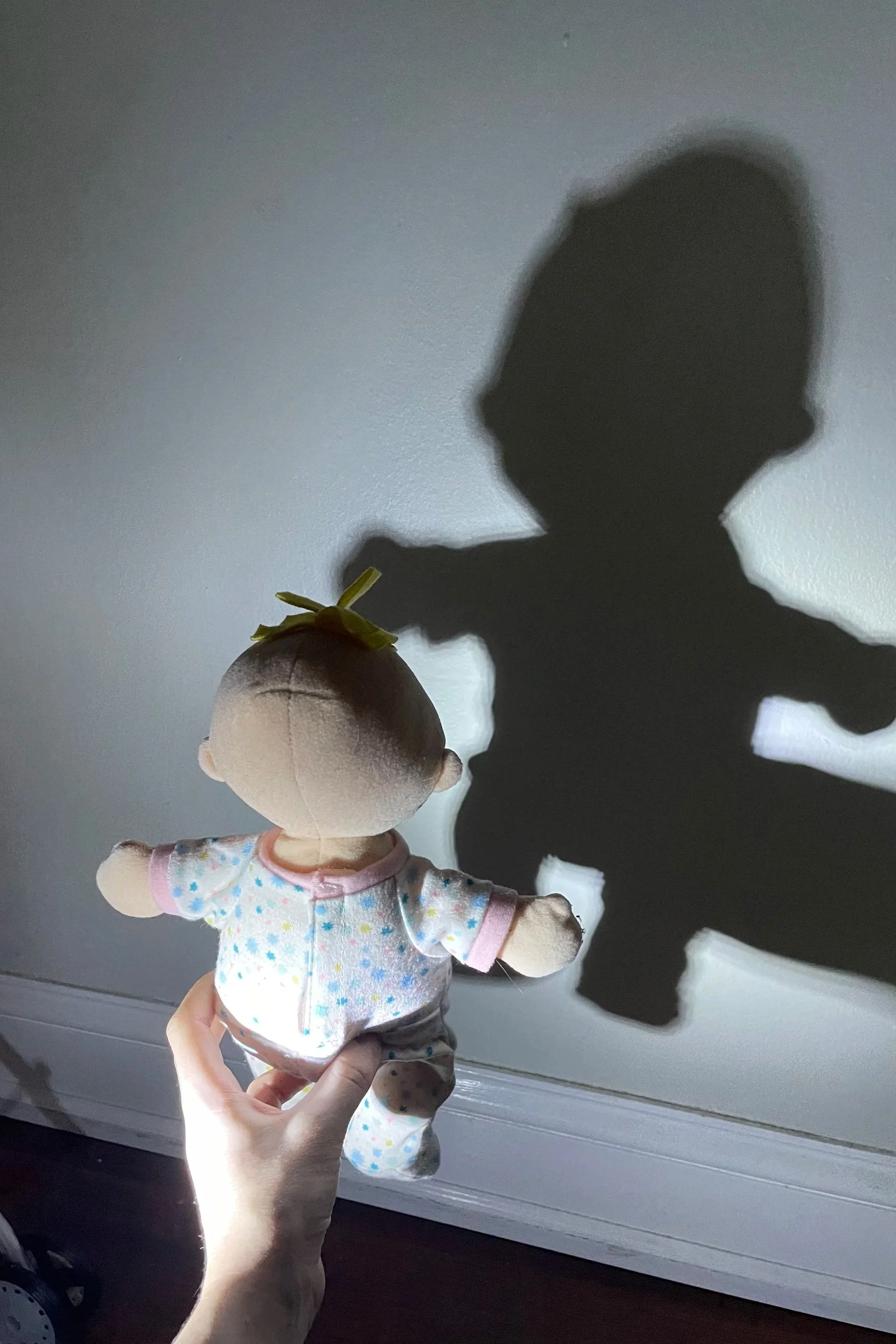 A plush doll with blonde hair, wearing a white shirt with colorful stars, held up by a person's hand, casting a shadow resembling a person's head and shoulders on a gray wall.
