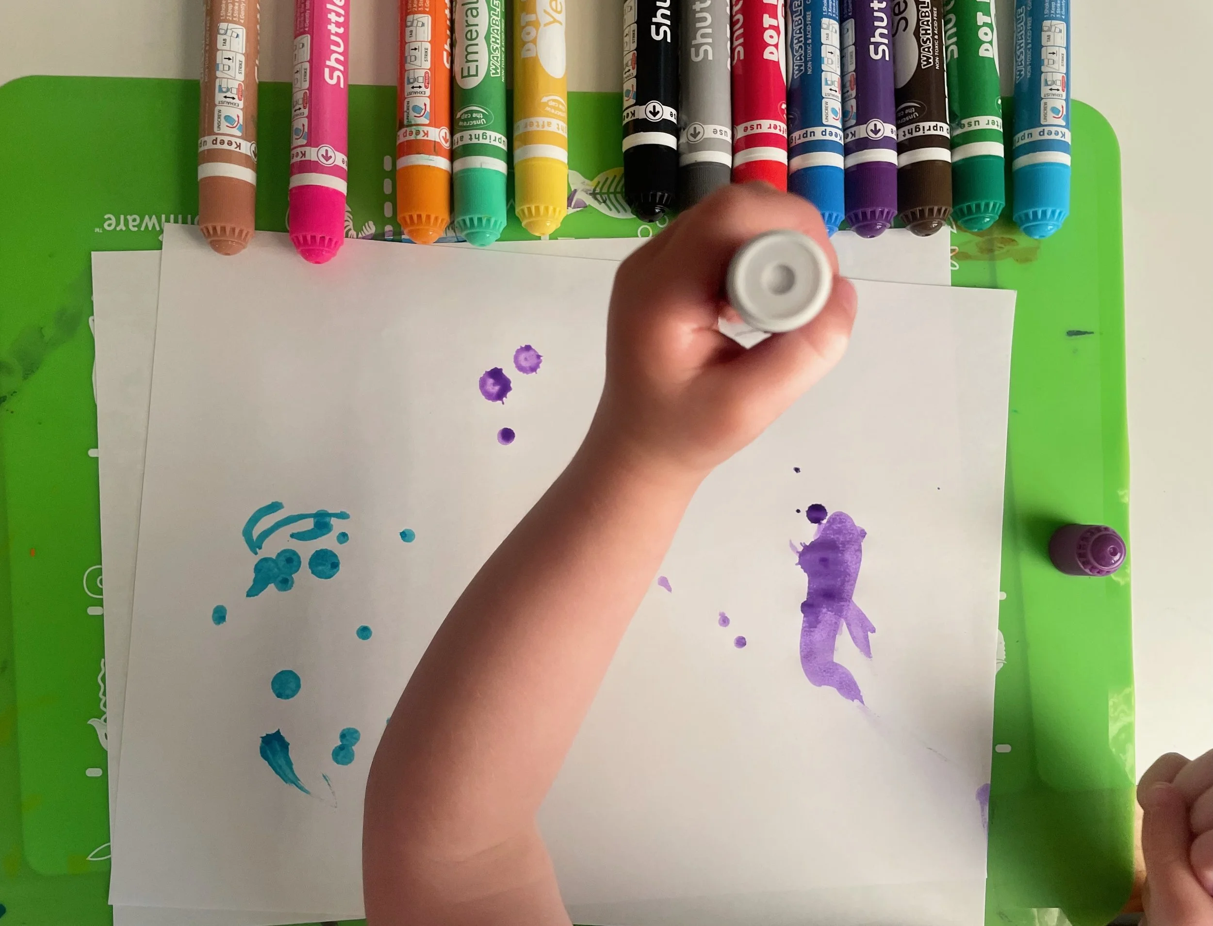 A hand holding a purple marker over a white sheet of paper with purple and blue watercolor paint splotches. Multiple colorful markers are lying horizontally on a green surface behind the paper.