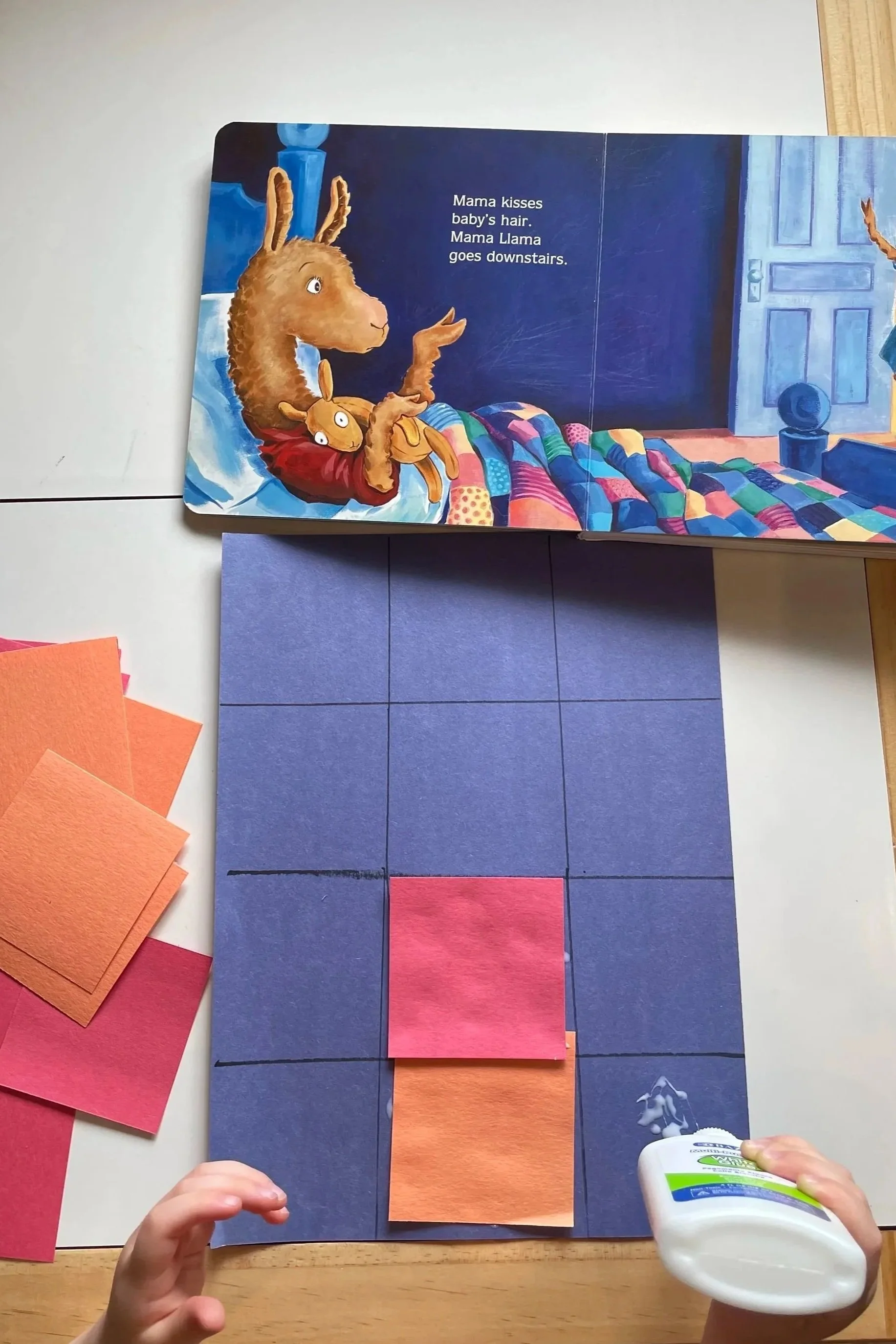 Image shows a child's craft project on a wooden table. The project includes a large dark blue paper rectangle, with orange and pink paper squares glued on it, mimicking a sunset or sunrise scene. There are also various colored paper squares and rectangles nearby, some in shades of orange, pink, and red. A hand in the bottom right corner holds a glue bottle, and another hand at the bottom left is reaching for one of the orange paper squares.