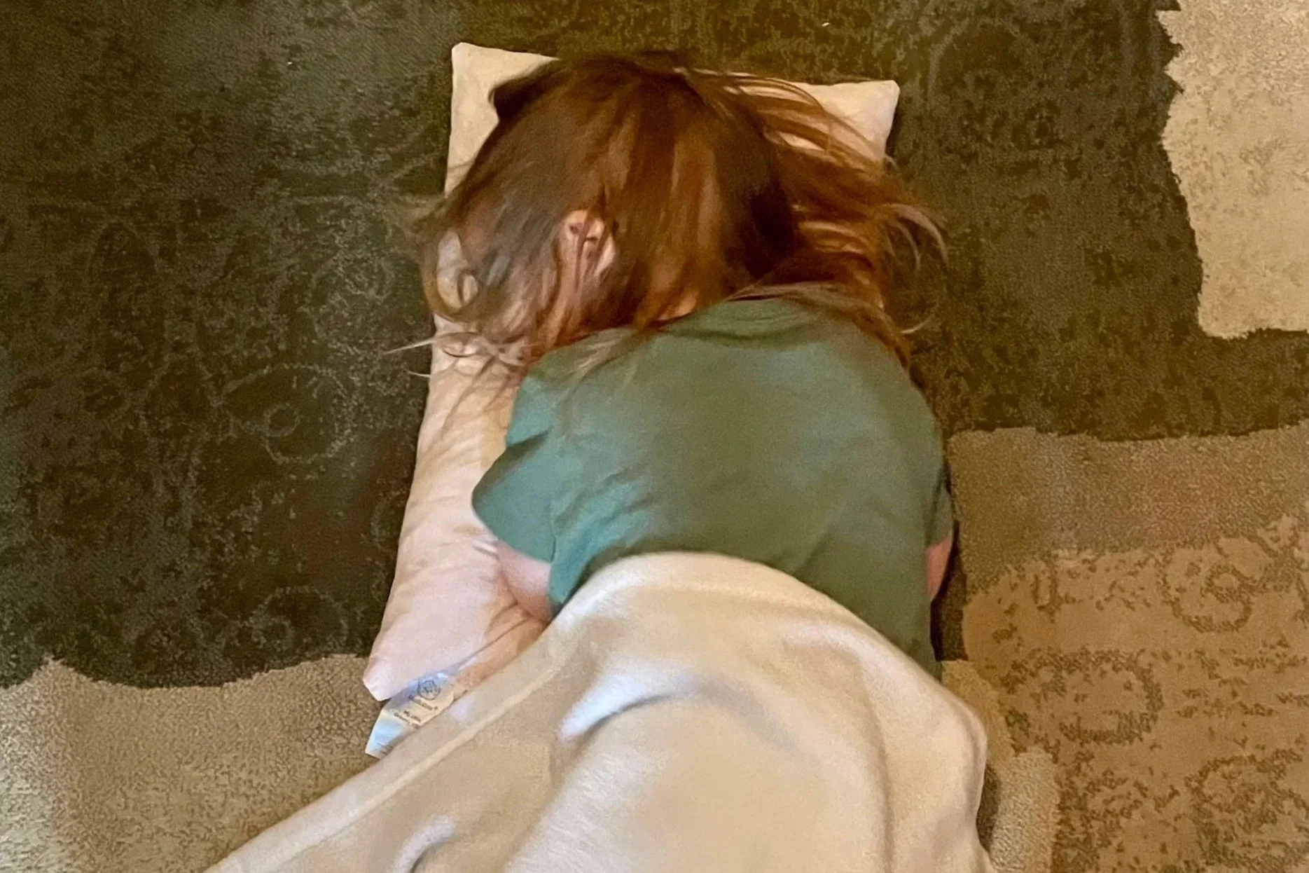 Person with brown hair lying face down on a pillow on the floor, with a carpet partially underneath, wearing a green shirt and white pants.