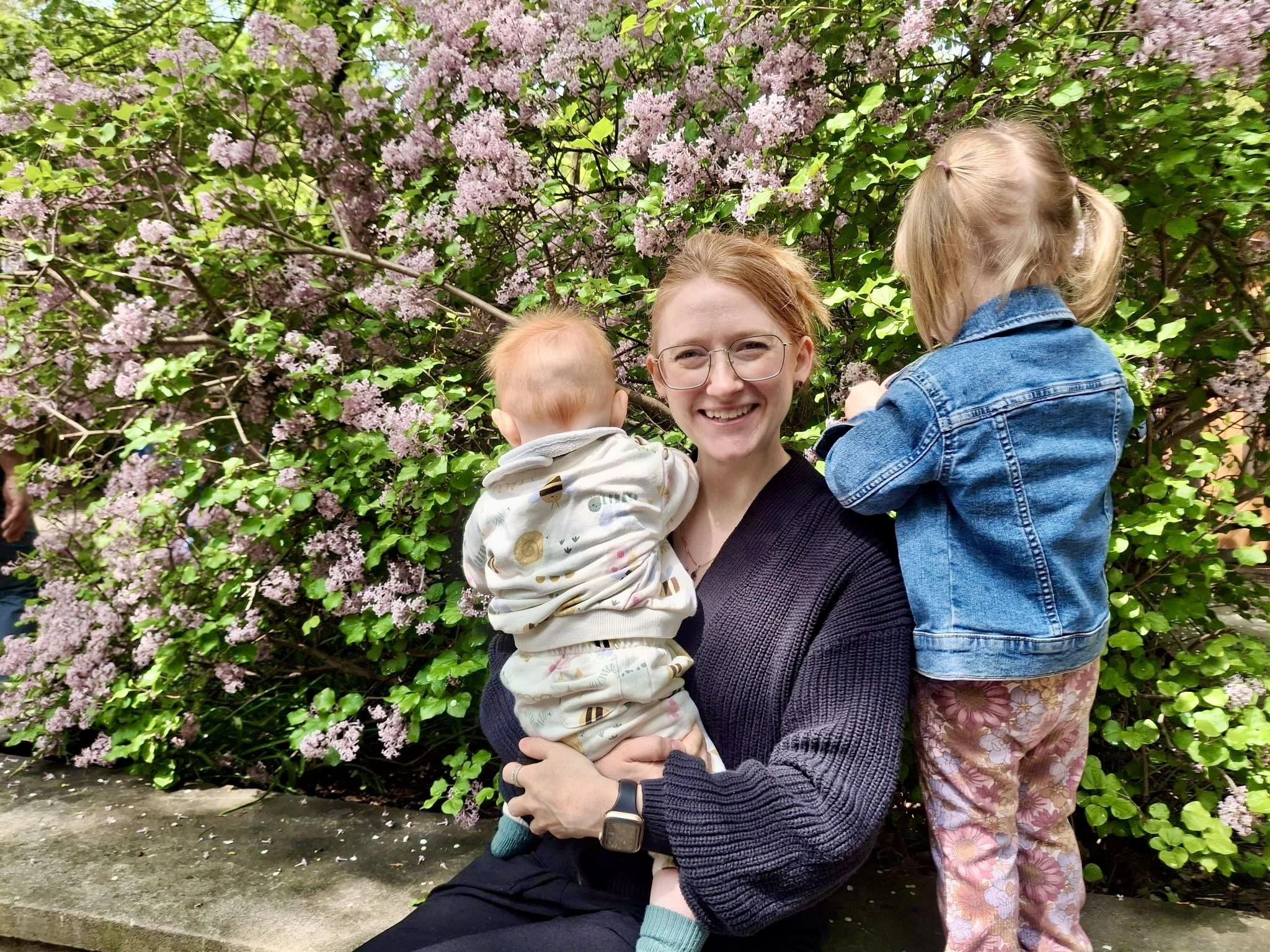 A woman with glasses holding a young child in front of a flowering bush with pink blossoms. Another child wearing a denim jacket is nearby, with only the back of their head and part of their body visible.