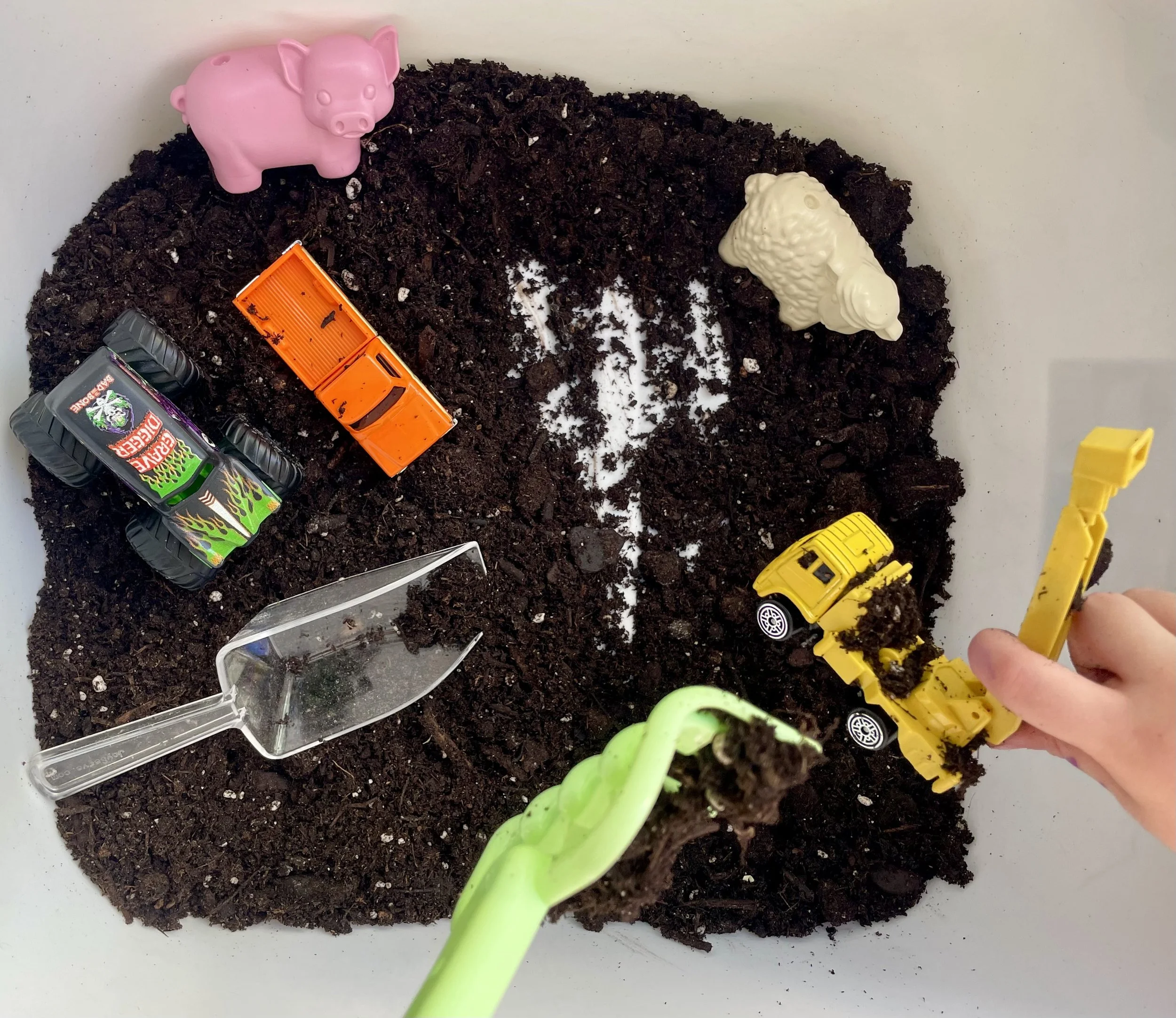 Children's toy cars and shovels placed on dark soil inside a white container.