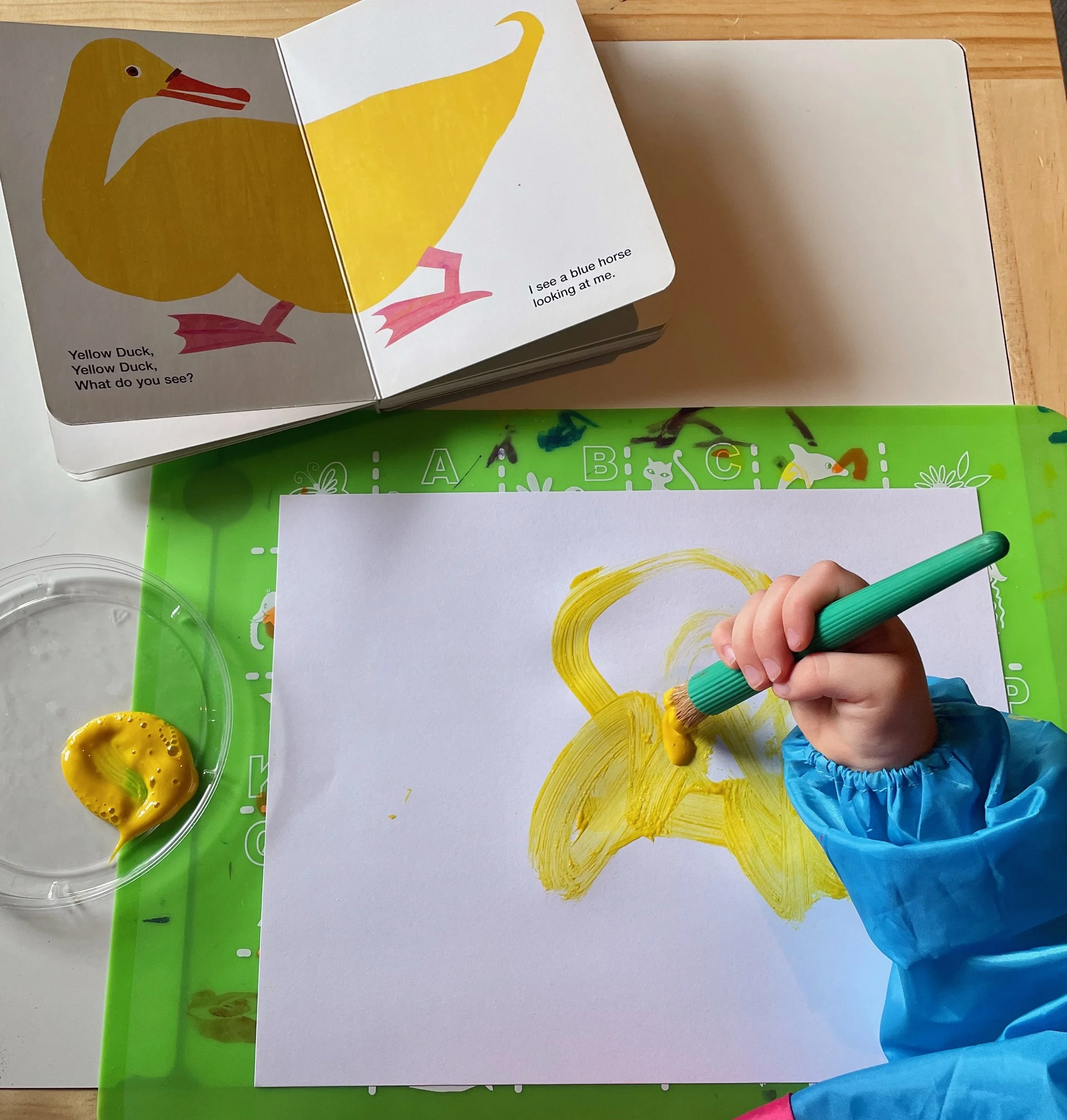A child's hand holding a green marker drawing a yellow shape on a white sheet of paper. The background shows a green educational play mat and a clear plastic bowl with a yellow rubber duck. The drawing is a large yellow arc, and the child is wearing a blue sleeve.