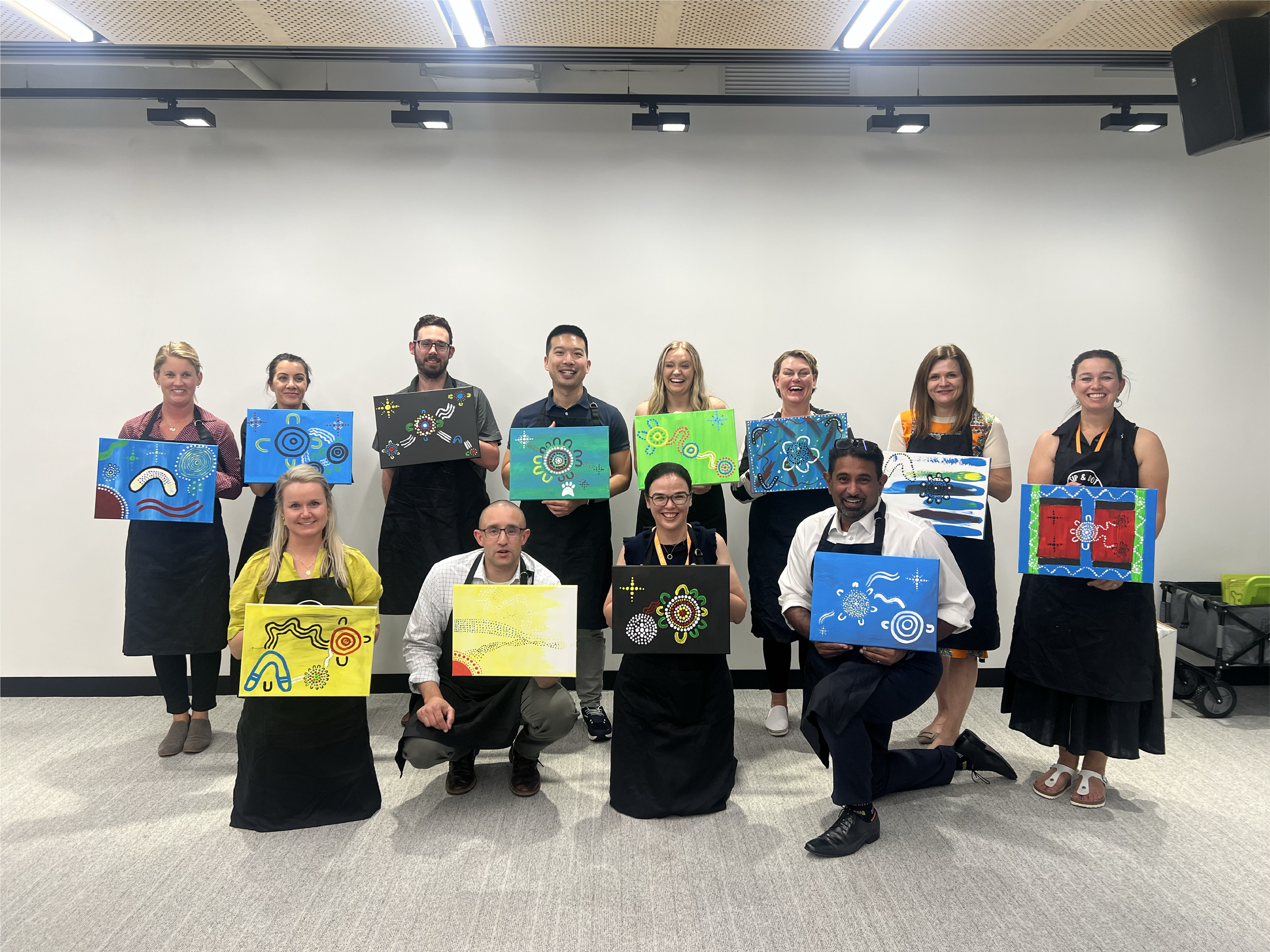 Group of people holding colorful abstract paintings in an art gallery or classroom with a plain white wall backdrop.