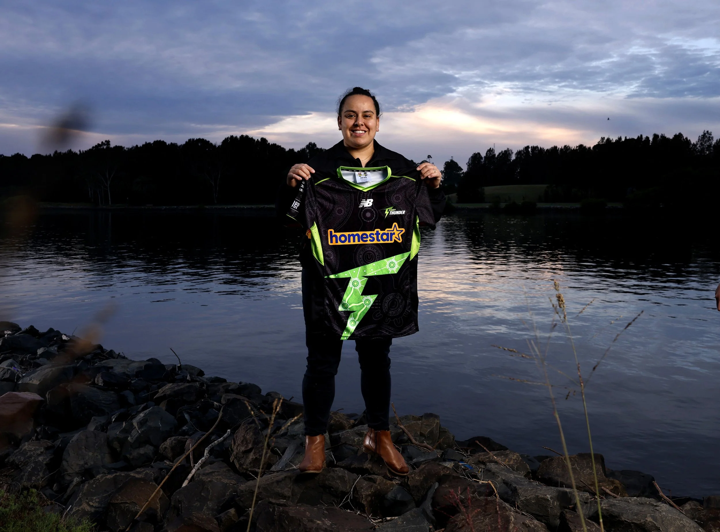 A woman standing on a rocky shore by a body of water during sunset, holding a sports jersey with the number 7 and the words 'homestar' on it, smiling at the camera.