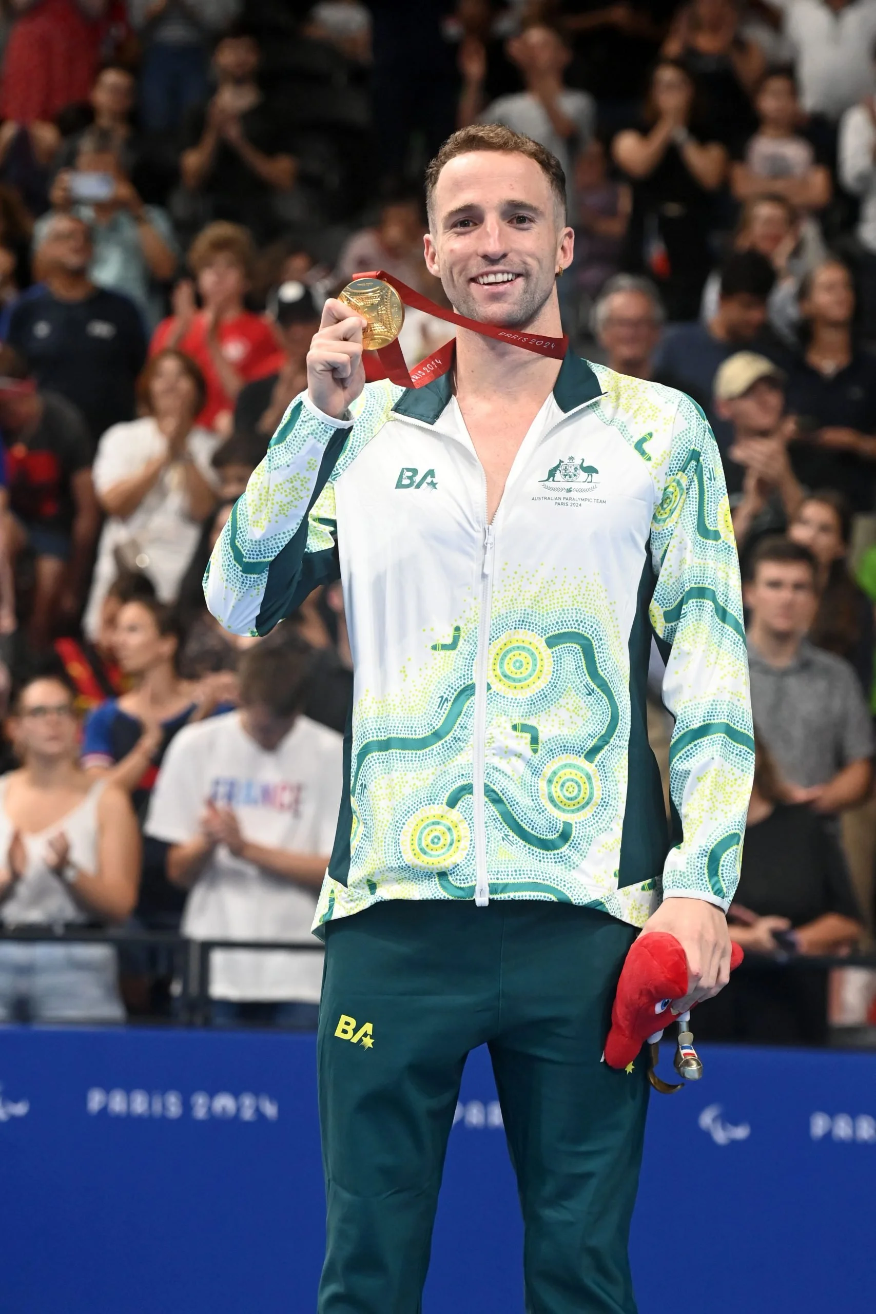 A male athlete from the Australian Paralympic team holding a gold medal at the Paris 2024 Paralympic Games, standing in front of a crowd during the award ceremony.
