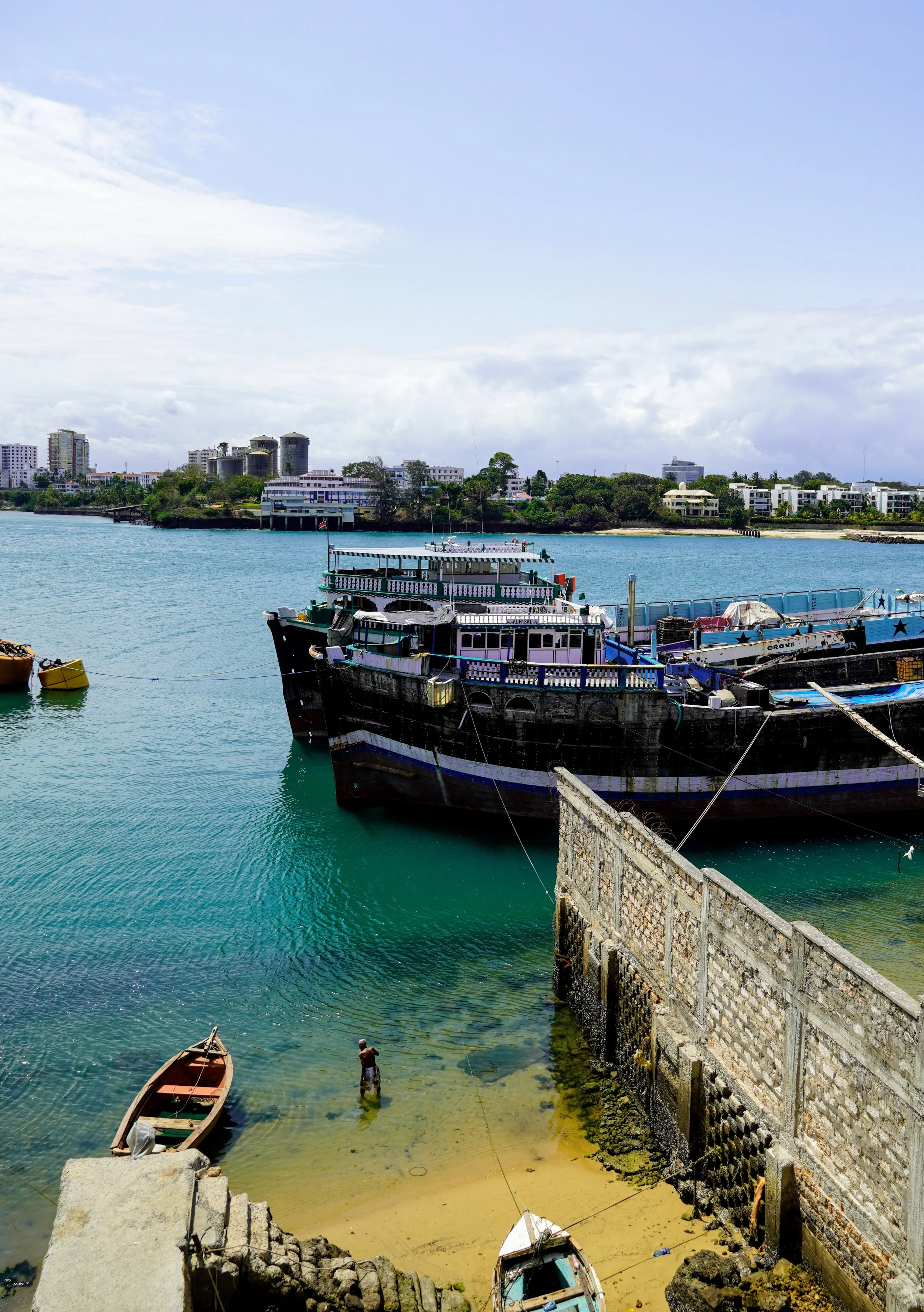 View of a small harbor with boats docked near a stone wall, a man standing in the shallow water, and a city skyline with buildings and trees in the background under a partly cloudy sky.
