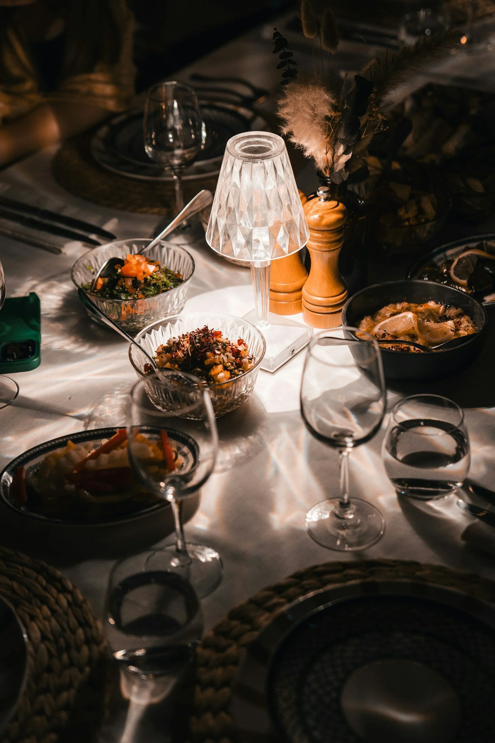 A table set with glassware, bowls of food, a lamp, and decorative items including wooden salt and pepper shakers and dried floral arrangements in a dimly lit environment.