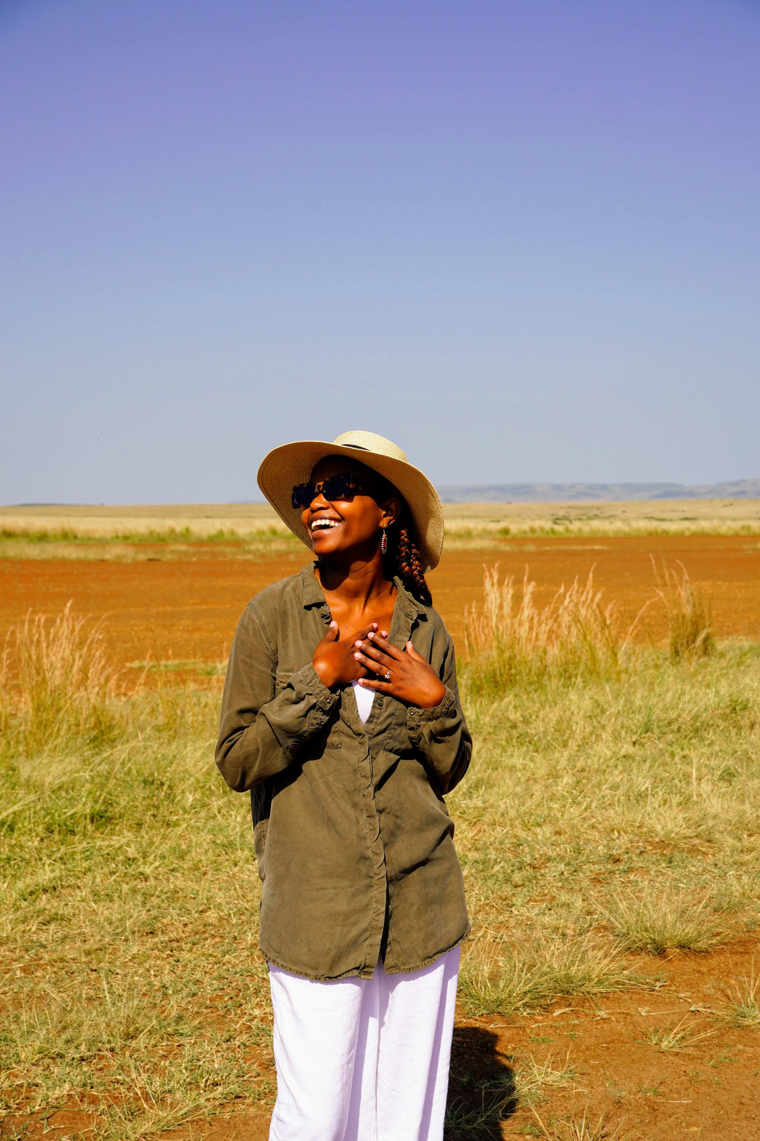 A woman standing outdoors in a grassy field under a clear blue sky, wearing a wide-brimmed hat, sunglasses, a green jacket, and white pants, smiling and touching her chest.