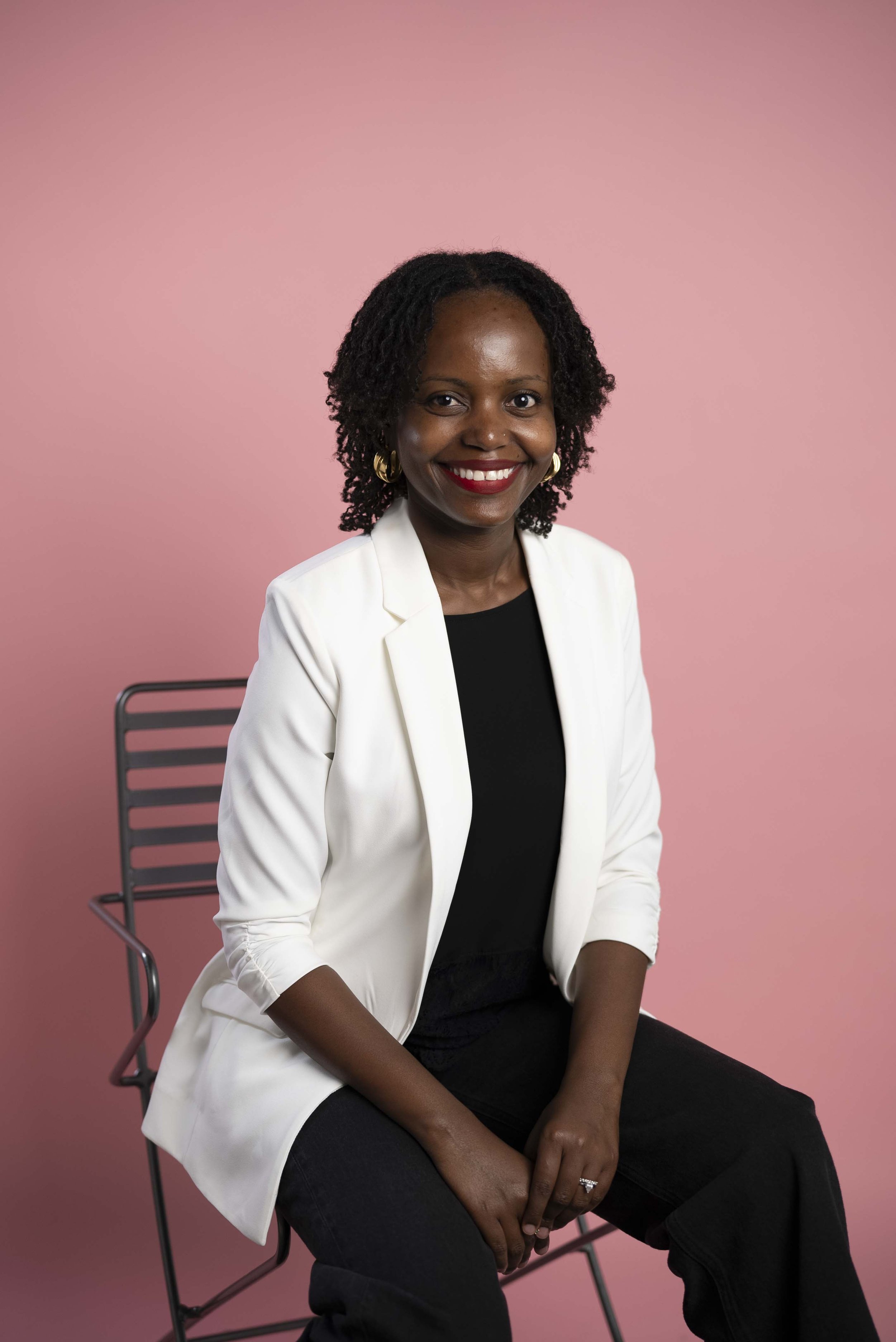 Smiling African American woman with curly hair, wearing a white blazer and black top, sitting on a metal chair against a pink background.