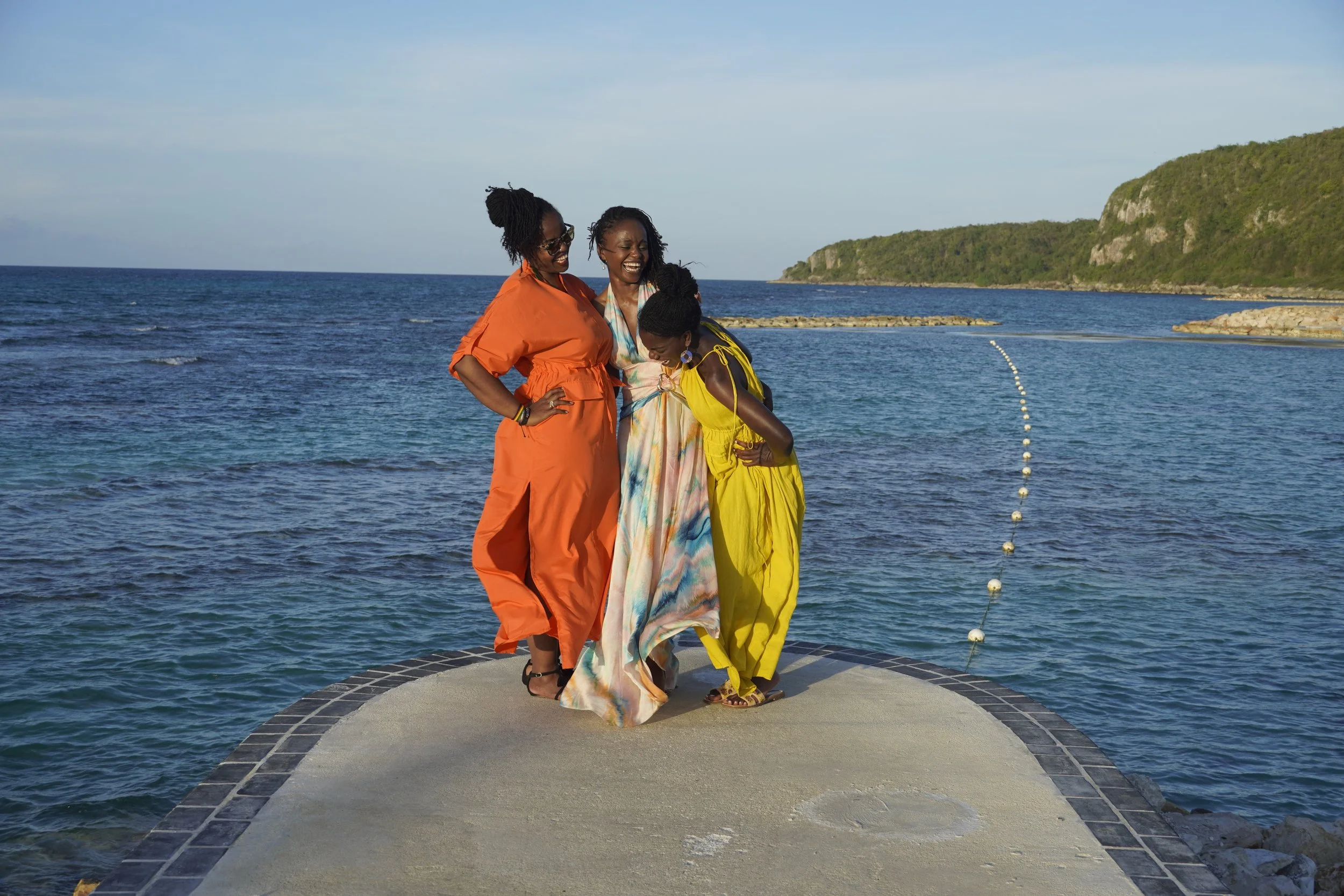Three women in colorful dresses are standing on a small circular walkway by the ocean, smiling and laughing, with a hilly green landscape in the background.