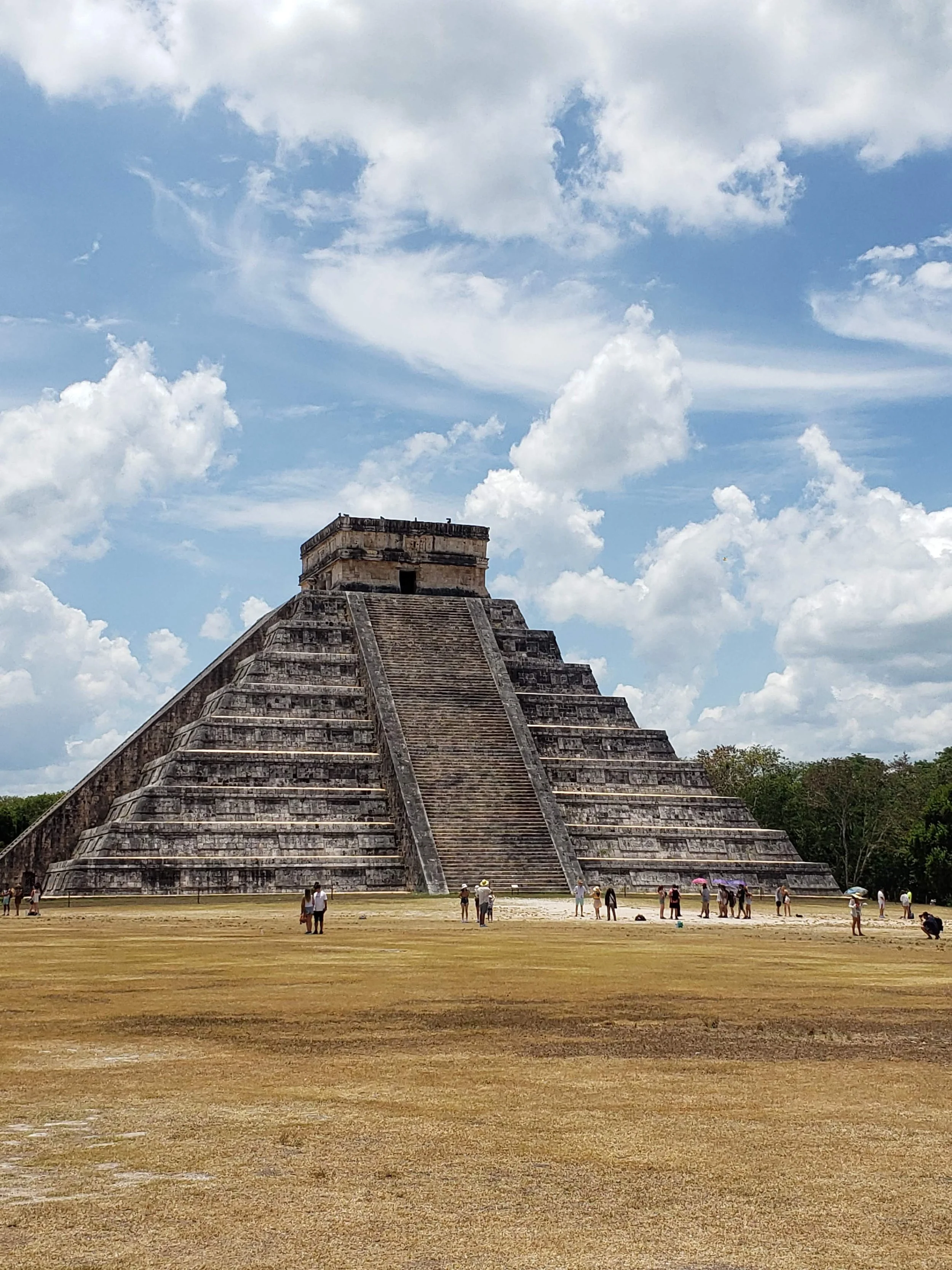 Ancient Mayan step pyramid at Chichen Itza, with tourists walking and taking photos, under a partly cloudy sky.