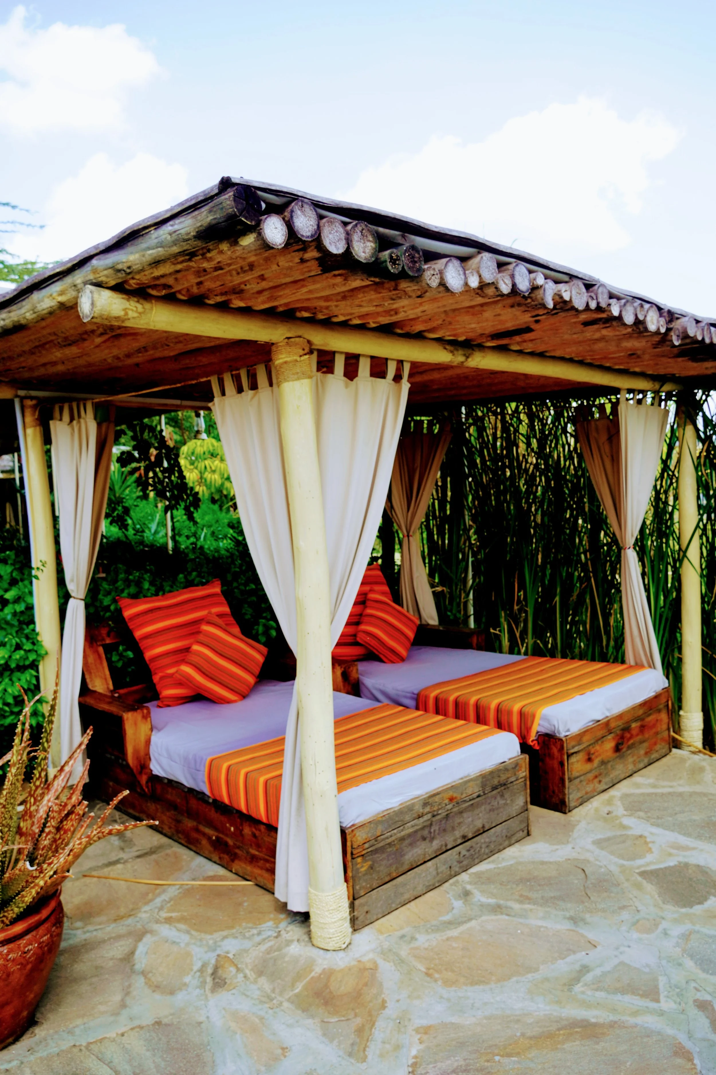 A wooden outdoor cabana with a thatched roof, white curtains, and striped cushions on a daybed, surrounded by tropical plants and overlooking a blue sky.