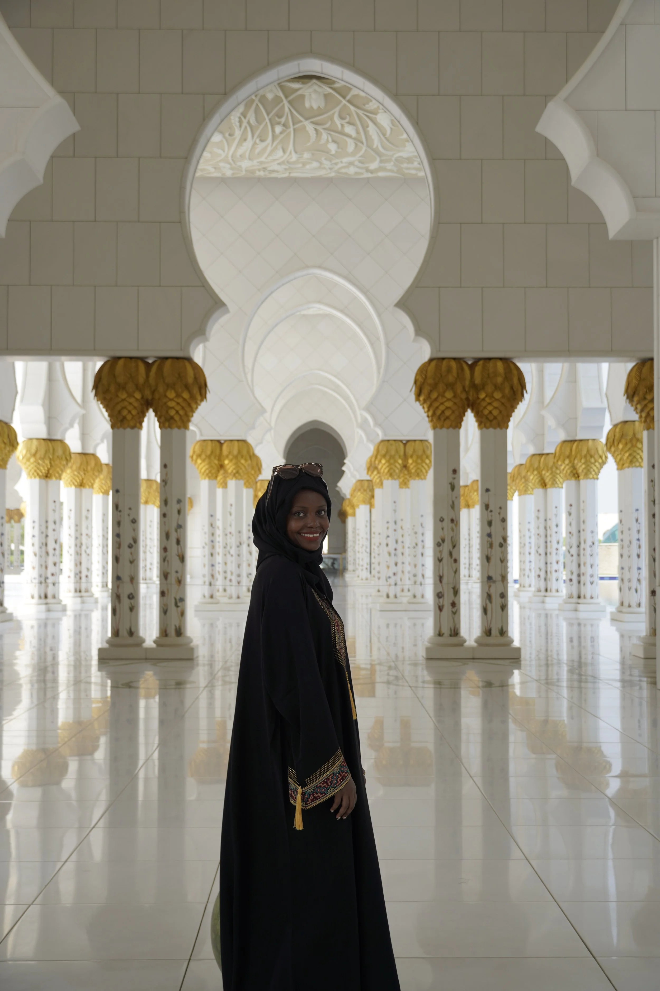 A woman in traditional attire and a black headscarf smiles in a white, ornate corridor with gold accents and multiple arches.