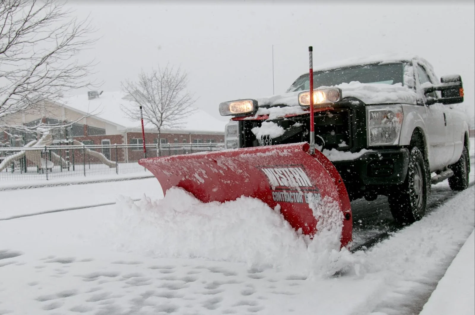 A pickup truck equipped with a snow plow blade clearing snow from a street during a snowstorm. The scene appears to be in a residential area with trees and houses in the background.