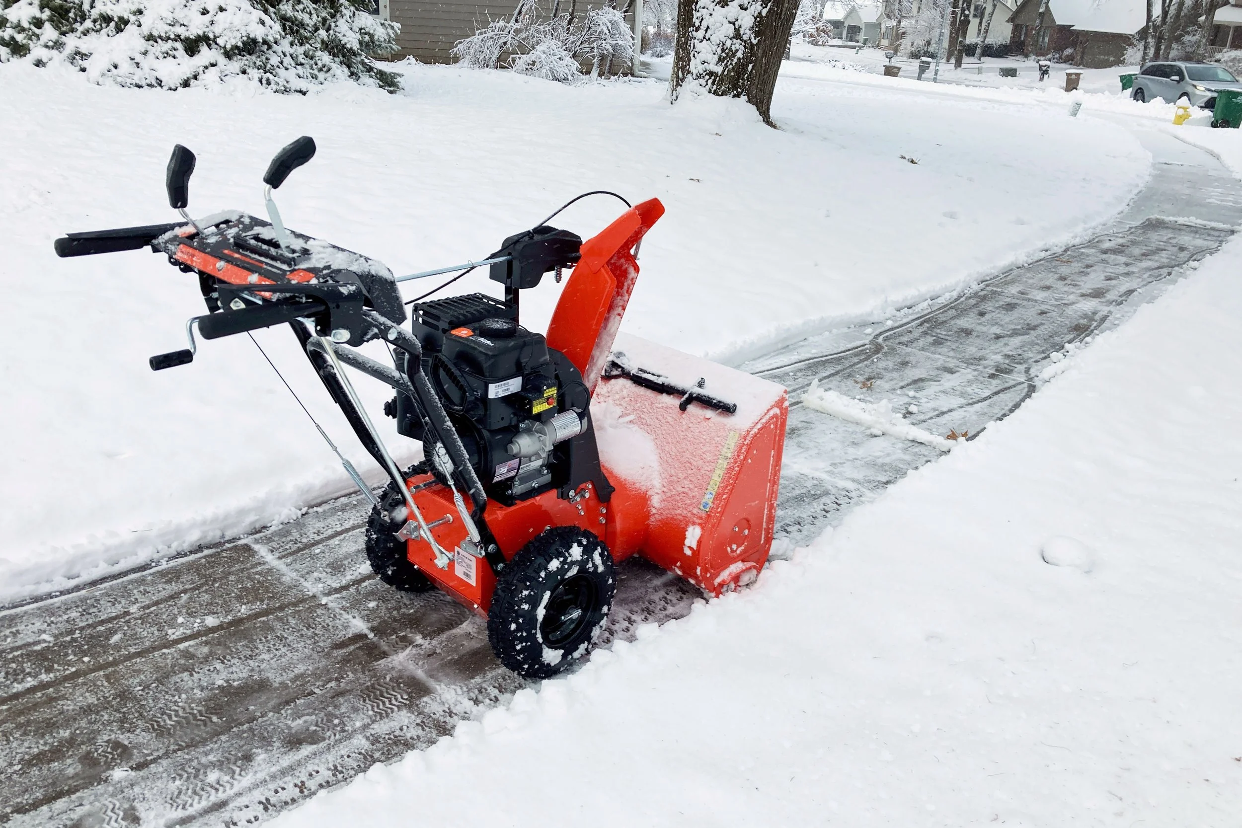 Snow blower clearing snow from a residential sidewalk on a snowy day, with snow-covered trees and houses in the background.