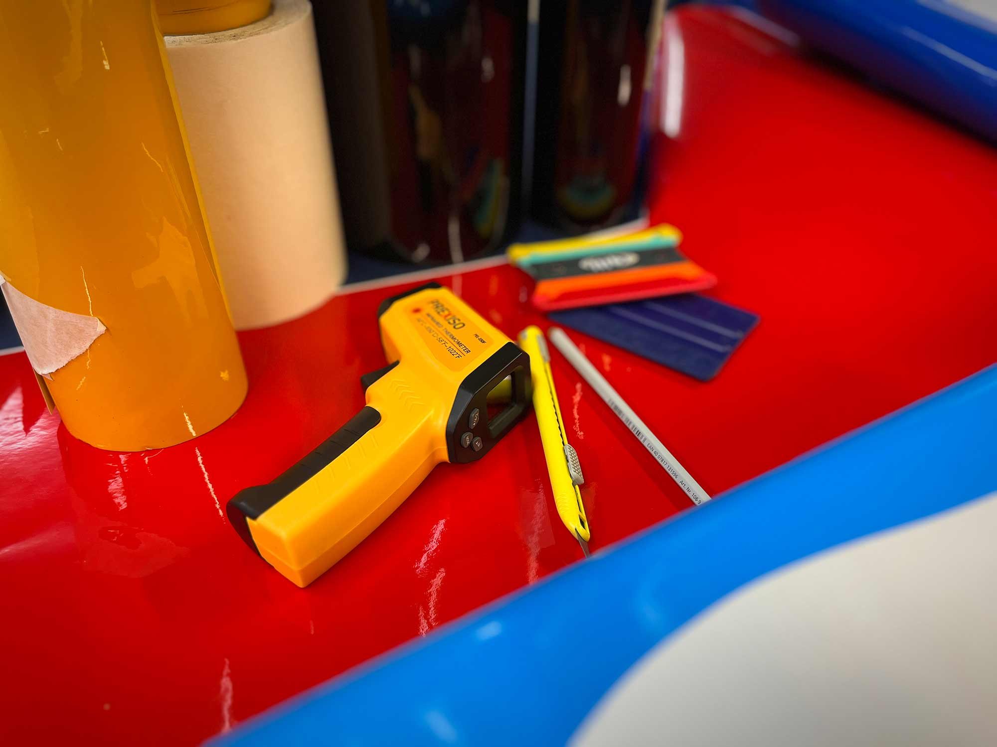 Close-up of a red surface with various school supplies including a yellow laser distance meter, yellow scissors, colored pencils, and folders, with a blue and yellow striped edge visible.