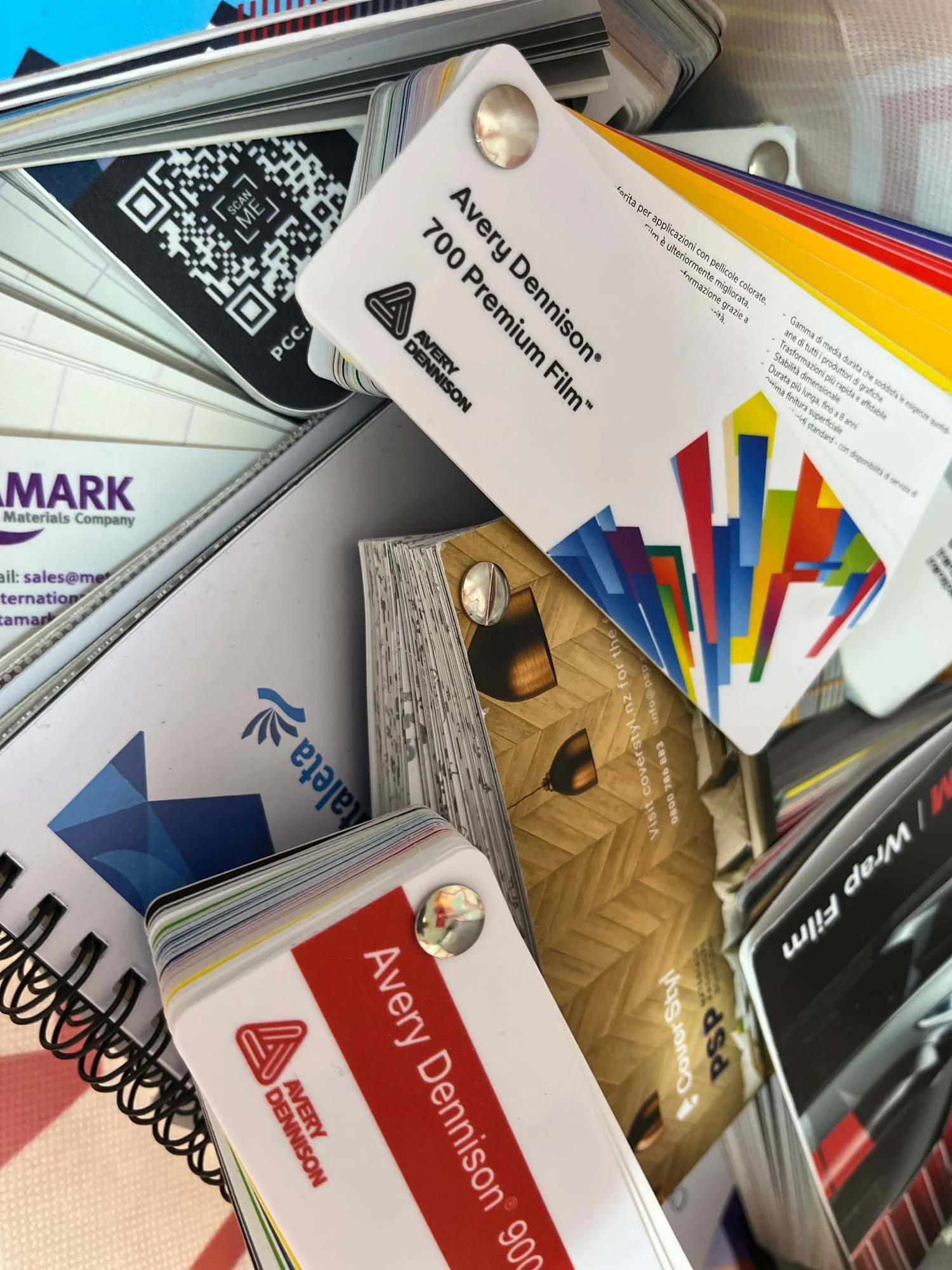 A display of various color swatch cards and sample books on a store shelf, including a white card with black text reading 'Avery Dennison 700 Premium Film' and other sample swatches with colorful stripes.