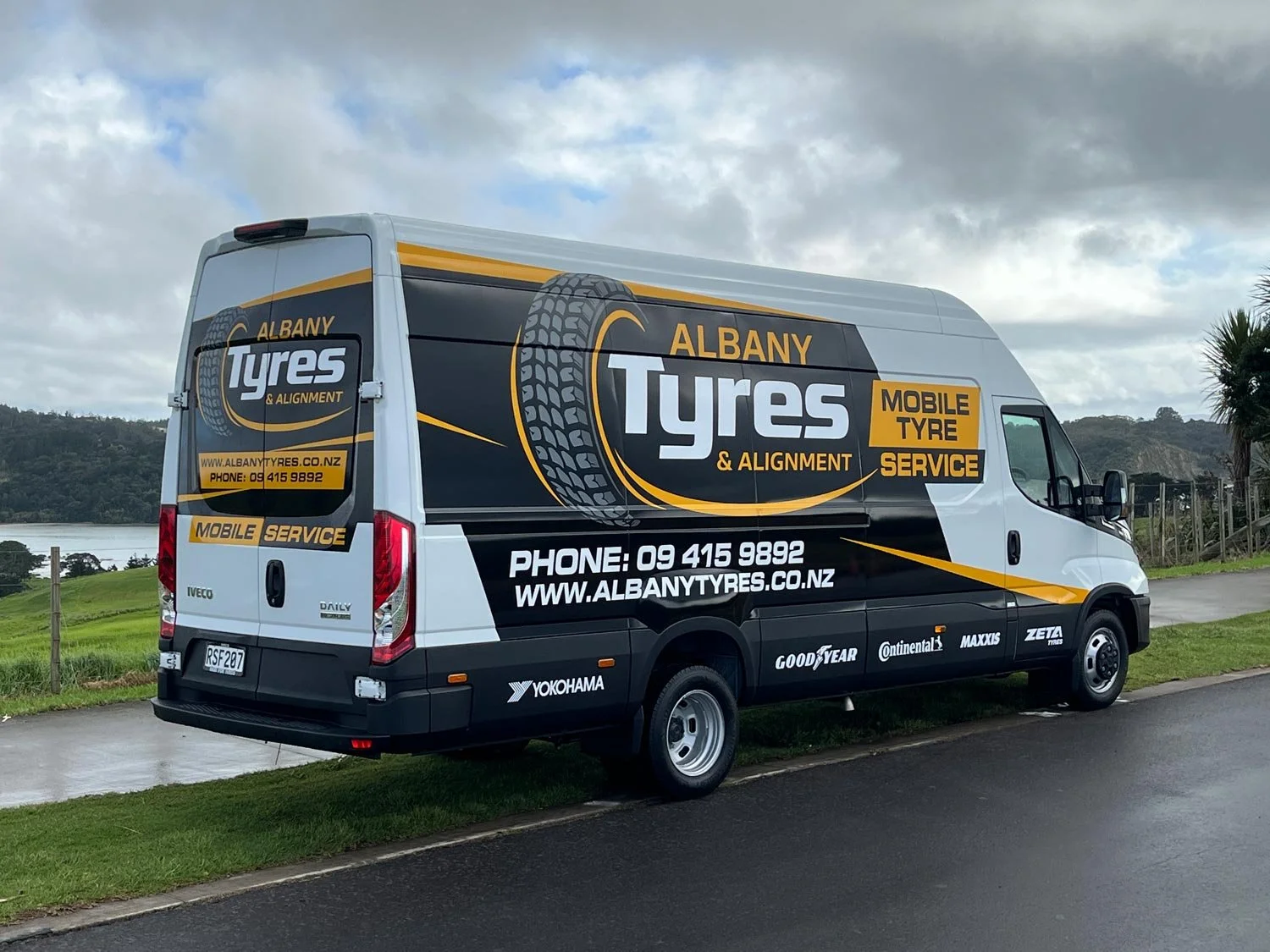 A white service van with black and yellow graphics advertising Albany Tyres, a mobile tyre and alignment service. The van displays the company’s website, phone number, and logos of tyre brands like Yokohama, Goodyear, Continental, Maxxis, and Zeta. The background features a cloudy sky, green grass, and a body of water.