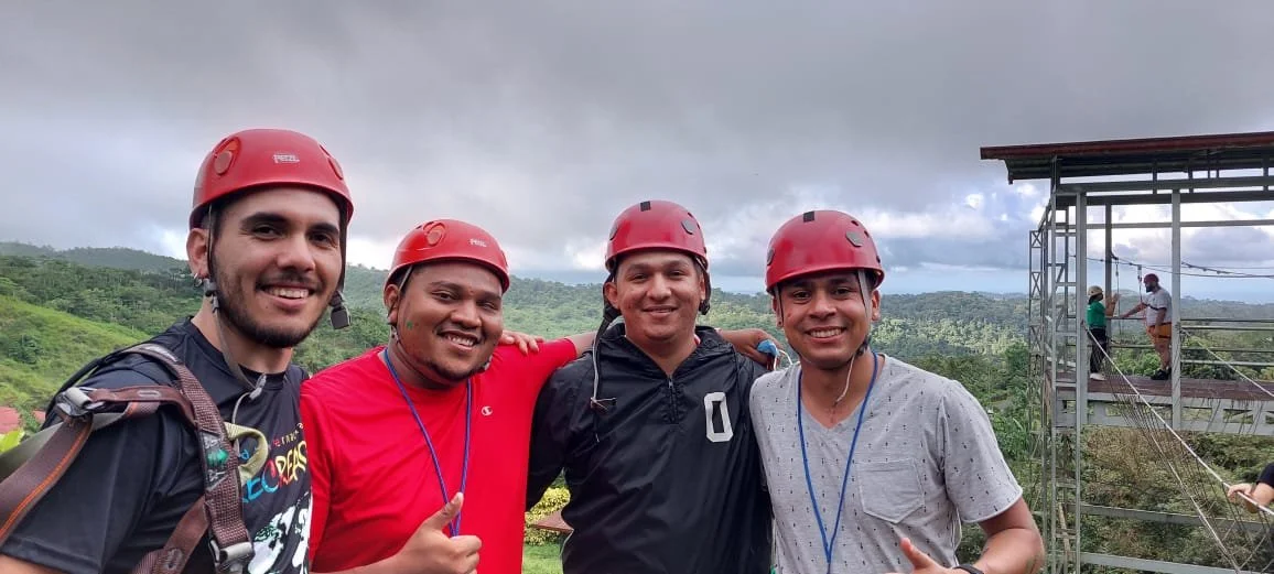 Cuatro hombres con casco de seguridad rosado en un lugar con naturaleza y una estructura de cuerdas en el fondo.