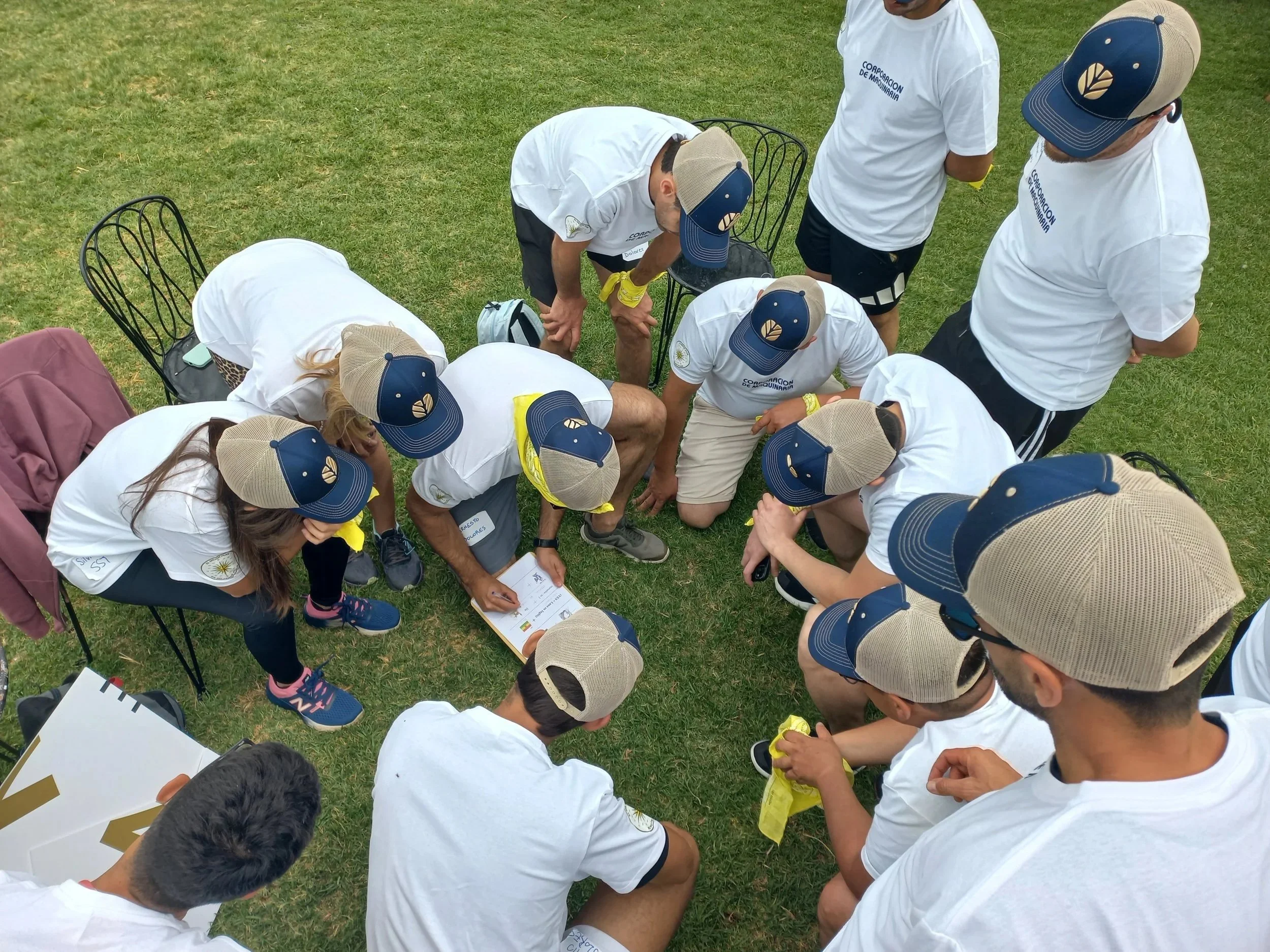 Grupo de personas con camisetas blancas, gorros con logo, y cintillas amarillas en un campo de césped participando en una actividad de equipo, revisando papeles y trabajando juntos.