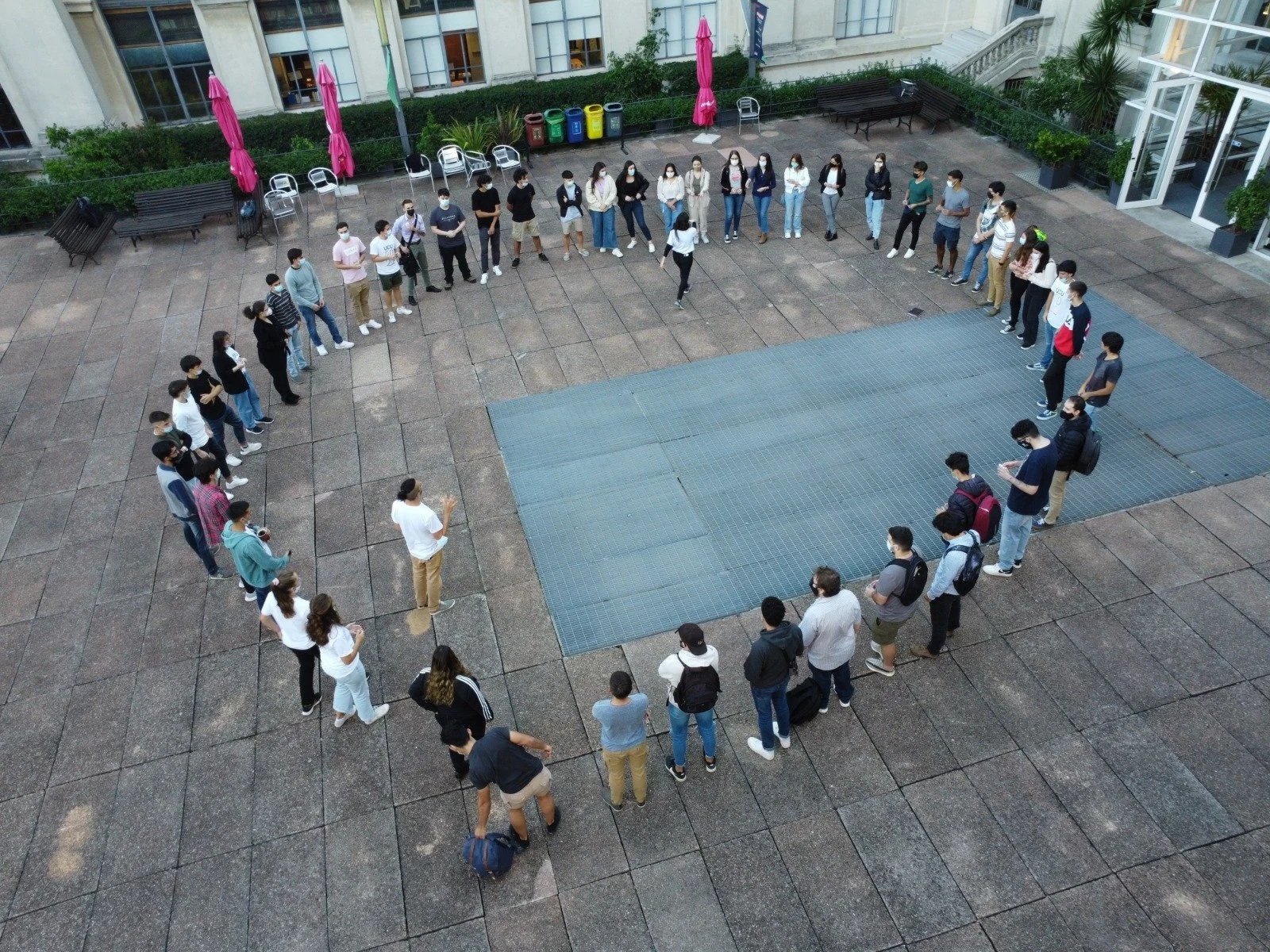 Grupo de personas usando mascarillas, formando un círculo en un espacio exterior de un edificio, algunas personas con mochilas, participando en una actividad grupal.