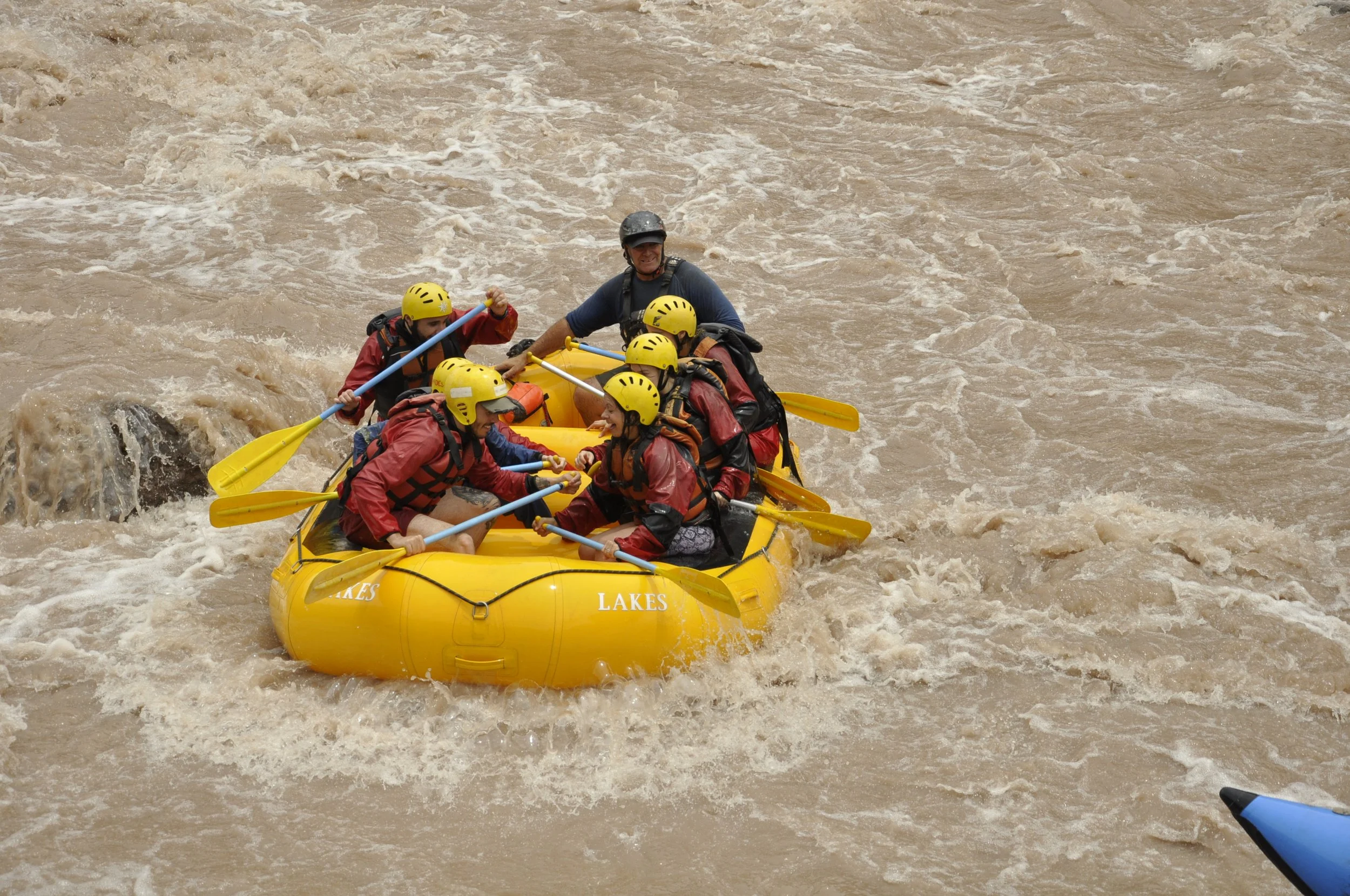 Grupo de personas haciendo rafting en aguas turbulentas con chalecos y cascos amarillos en un bote amarillo en un río de agua marrón.