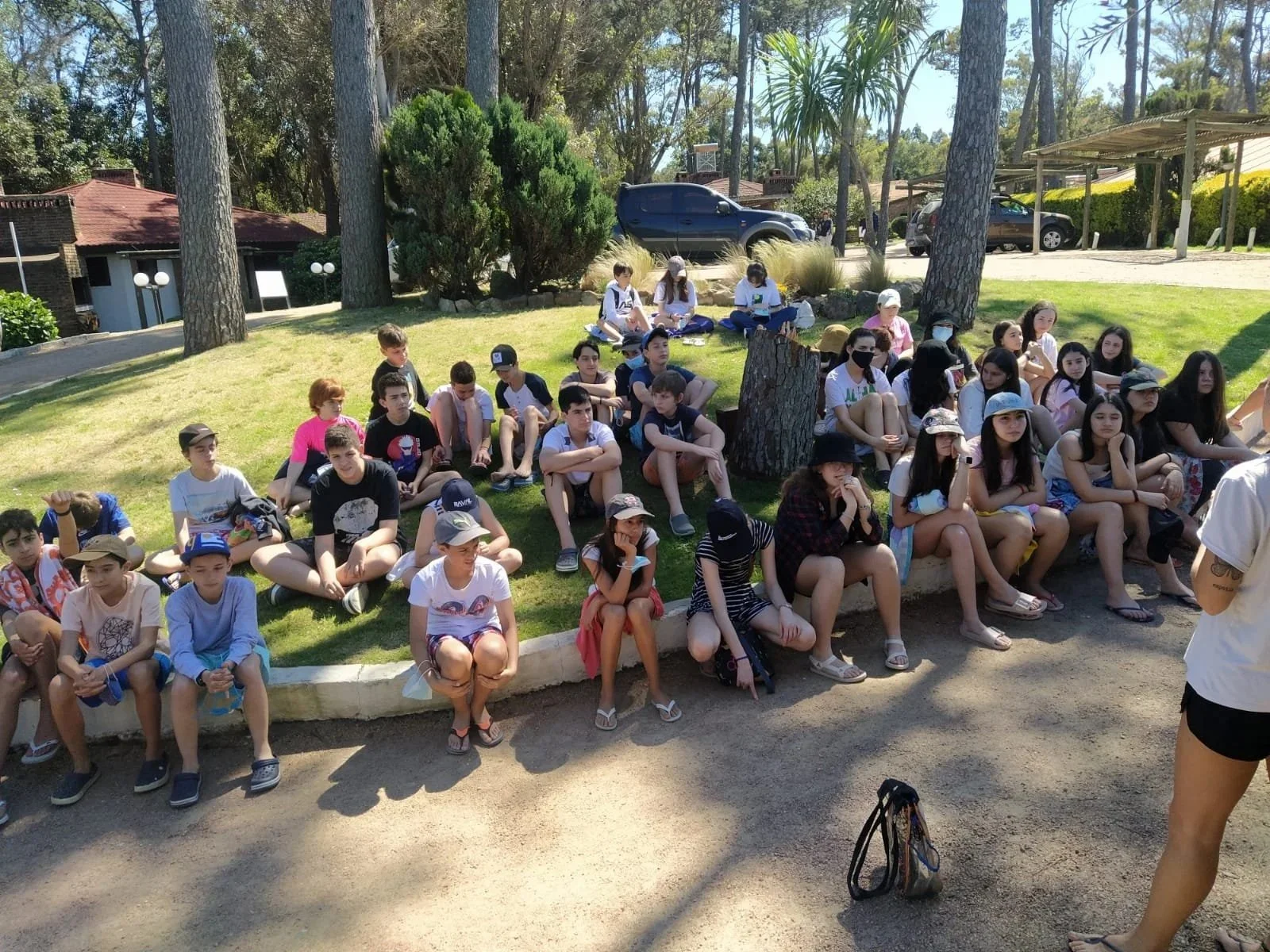 Grupo de niños y adolescentes sentados en un área ajardinada, algunos con gorra y máscara, en un ambiente al aire libre rodeado de árboles y vehículos estacionados.