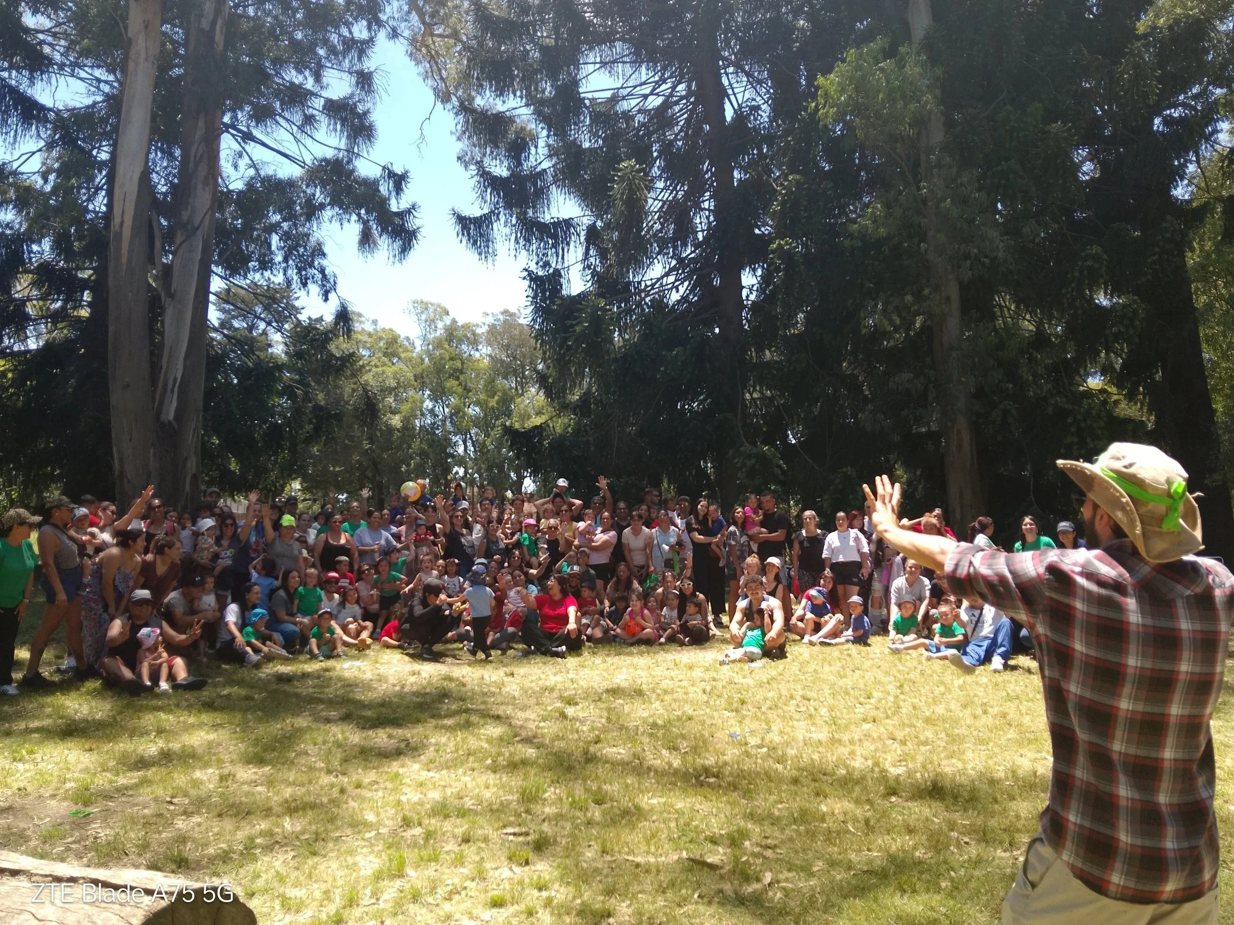 Grupo de personas, adultos y niños, reunidos en un parque rodeados de árboles, algunos niños con gorros y todos parecen participar en un evento o actividad group rally.