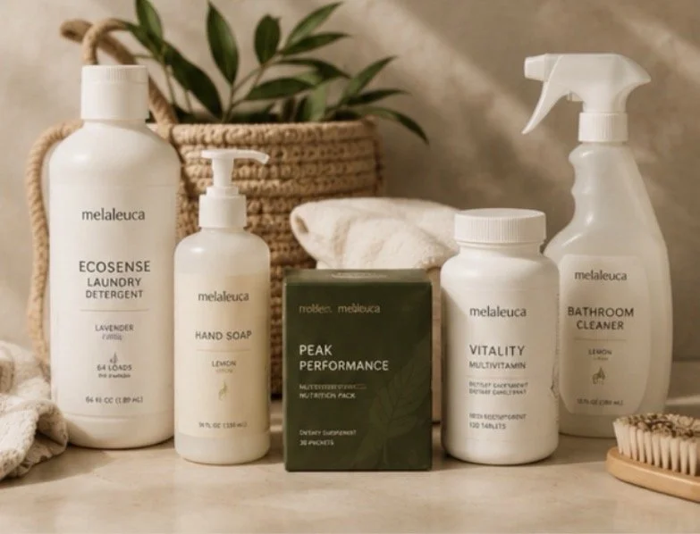 A collection of personal care and cleaning products on a countertop, including laundry detergent, hand soap, a dietary supplement, multivitamins, bathroom cleaner, and a container of brushes, with a woven basket, towel, greenery, and a wooden tray in the background.
