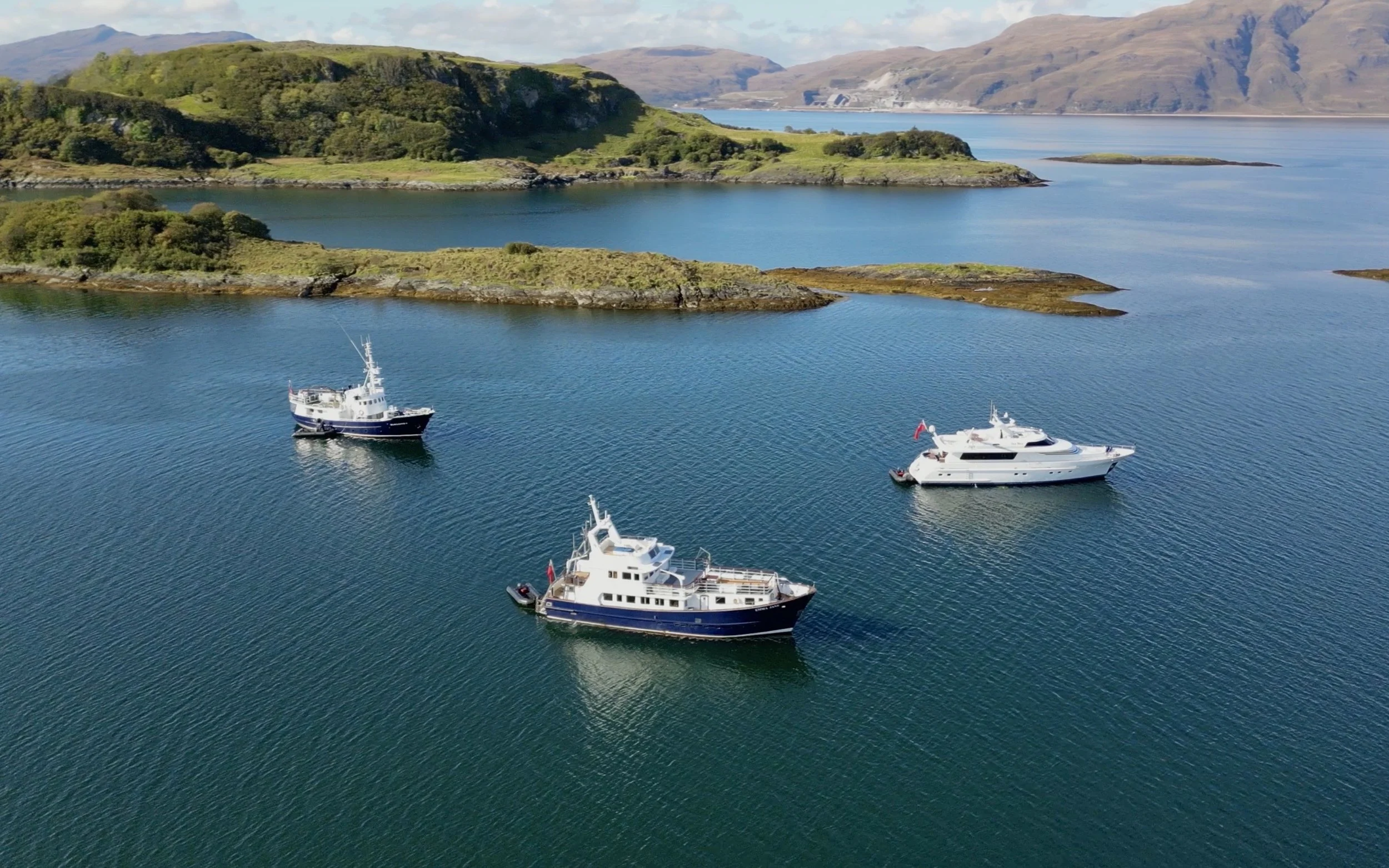 Aerial view of boats anchored in a calm body of water near green hilly islands with mountains in the background.