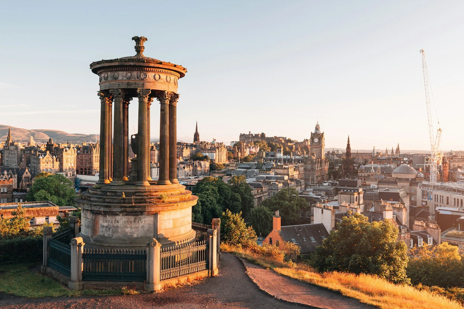 A historic monument with columns and a dome, overlooking a cityscape of Edinburgh, Scotland, including Big Ben, with a clear sky and sunset lighting.